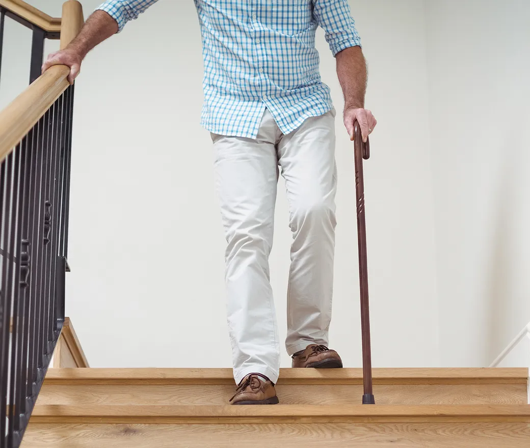 A man descending wooden home stairs, holding the handrail with one hand and a cane in the other