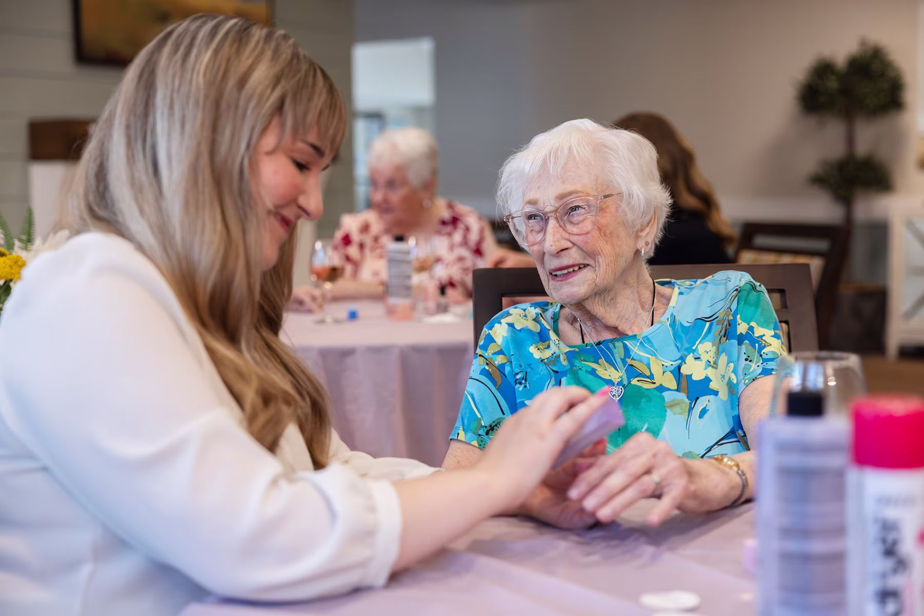 An elderly woman with white hair and glasses wearing a blue floral top smiles while holding hands with a younger woman with blonde hair in a white blouse at a table covered with a light pink tablecloth, with other people and tables blurred in the background.
