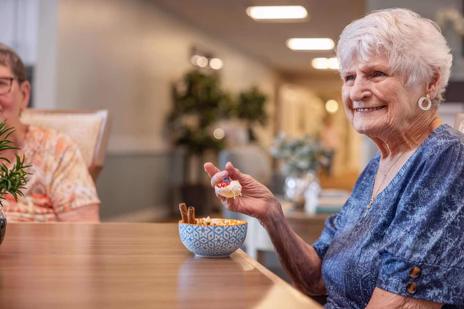An elderly woman with white hair wearing a blue shirt sits at a wooden table smiling and holding a small sandwich with cream and a cherry tomato. Another older woman is partially visible beside her in a floral shirt.
