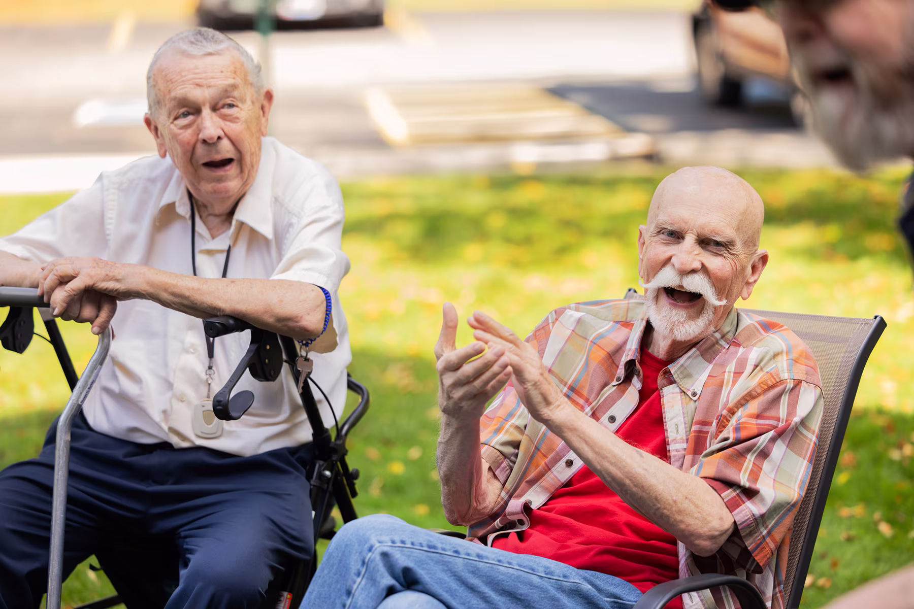 Two elderly men outdoors in a park setting, one sitting in a wheelchair listening attentively, and the other in a chair smiling and gesturing with his hands during a conversation.