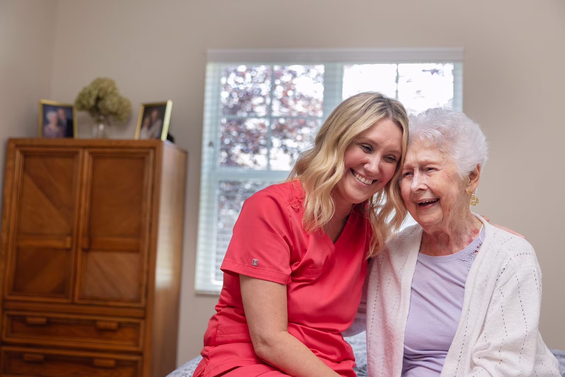A smiling caregiver in a red uniform embracing and sitting closely with a happy elderly woman wearing a white cardigan and lavender top inside a warmly lit room with a window and wooden furniture in the background.