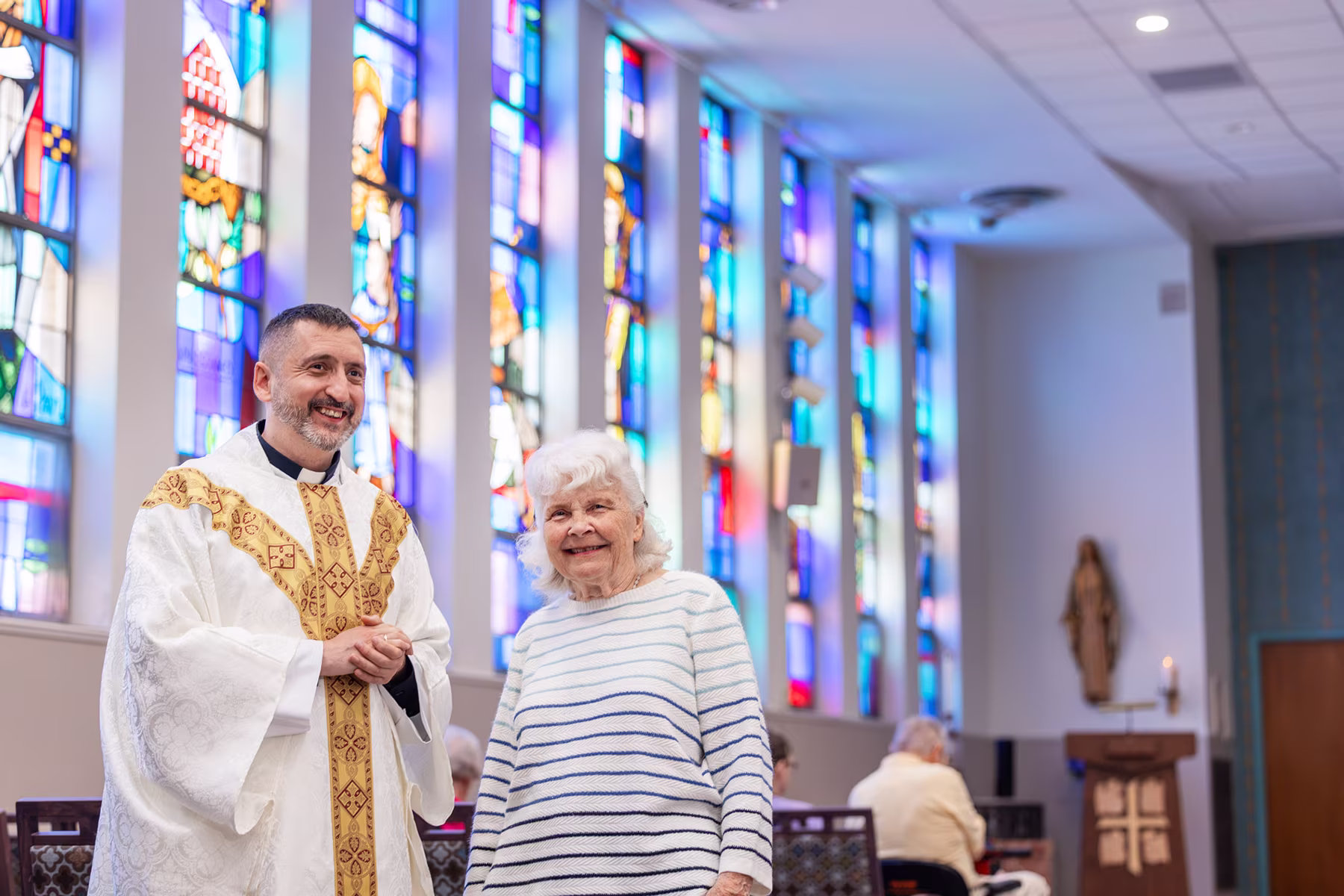 A smiling priest in white and gold vestments stands next to an elderly woman wearing a white sweater with blue stripes inside a church with colorful stained glass windows.