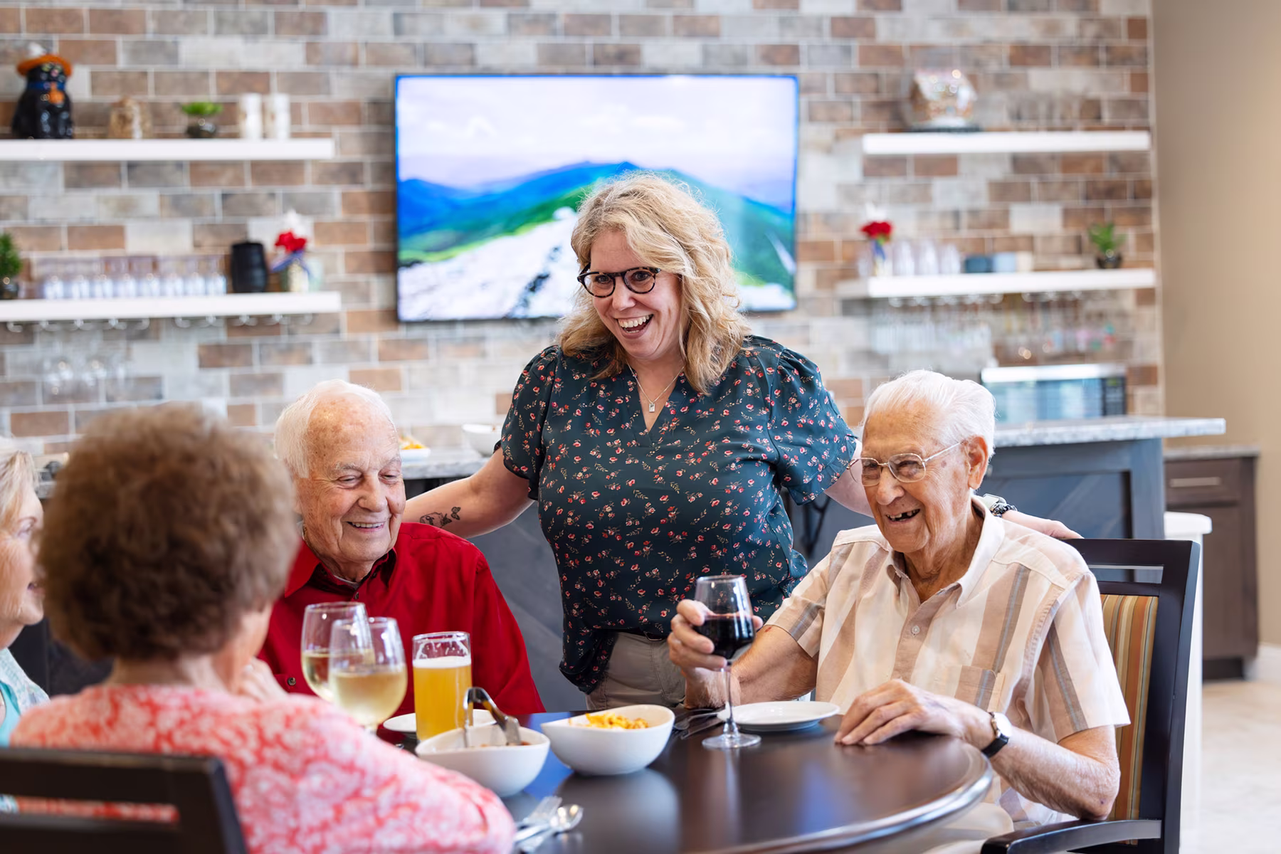A group of elderly people sitting around a table with drinks and snacks, smiling and enjoying social time with a younger woman standing behind them with her arms around two of the seated men in a bright room with a TV and shelves in the background.
