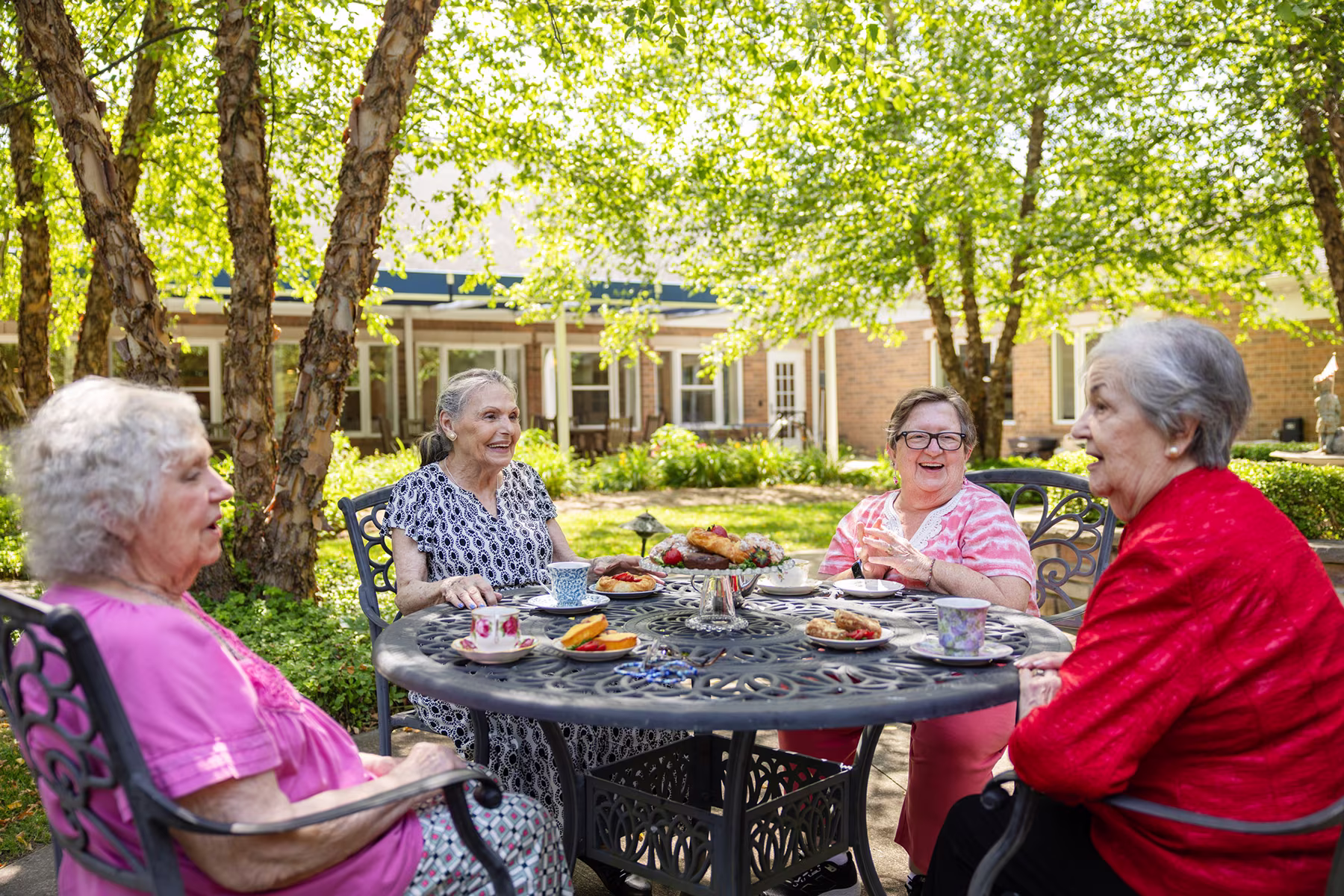 Four elderly women sitting around a black metal outdoor table enjoying tea and pastries in a sunny garden with trees and a building in the background.