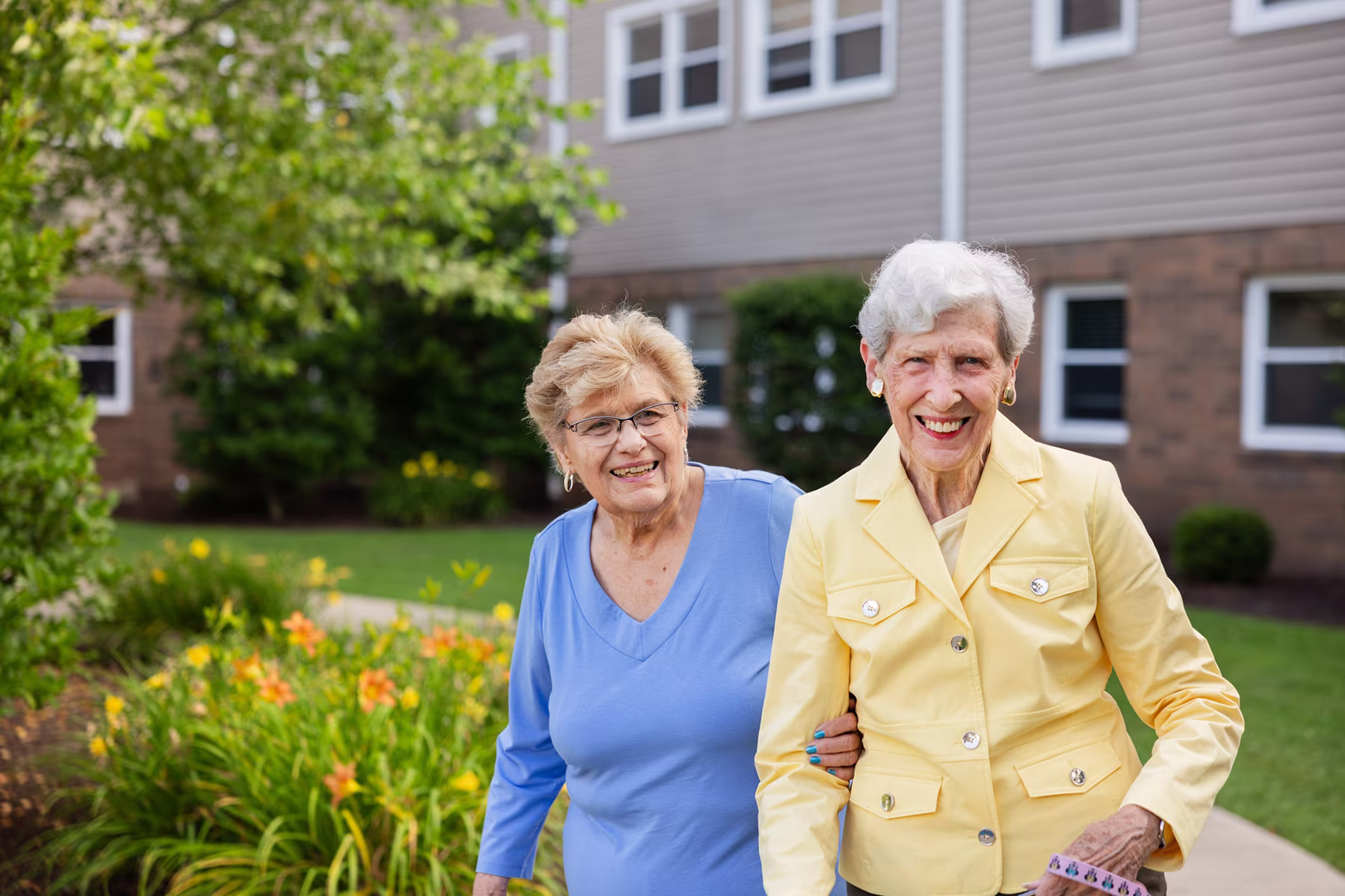 Two elderly women smiling outdoors near a building; one wearing a yellow jacket and the other in a blue top, standing close and holding each other.