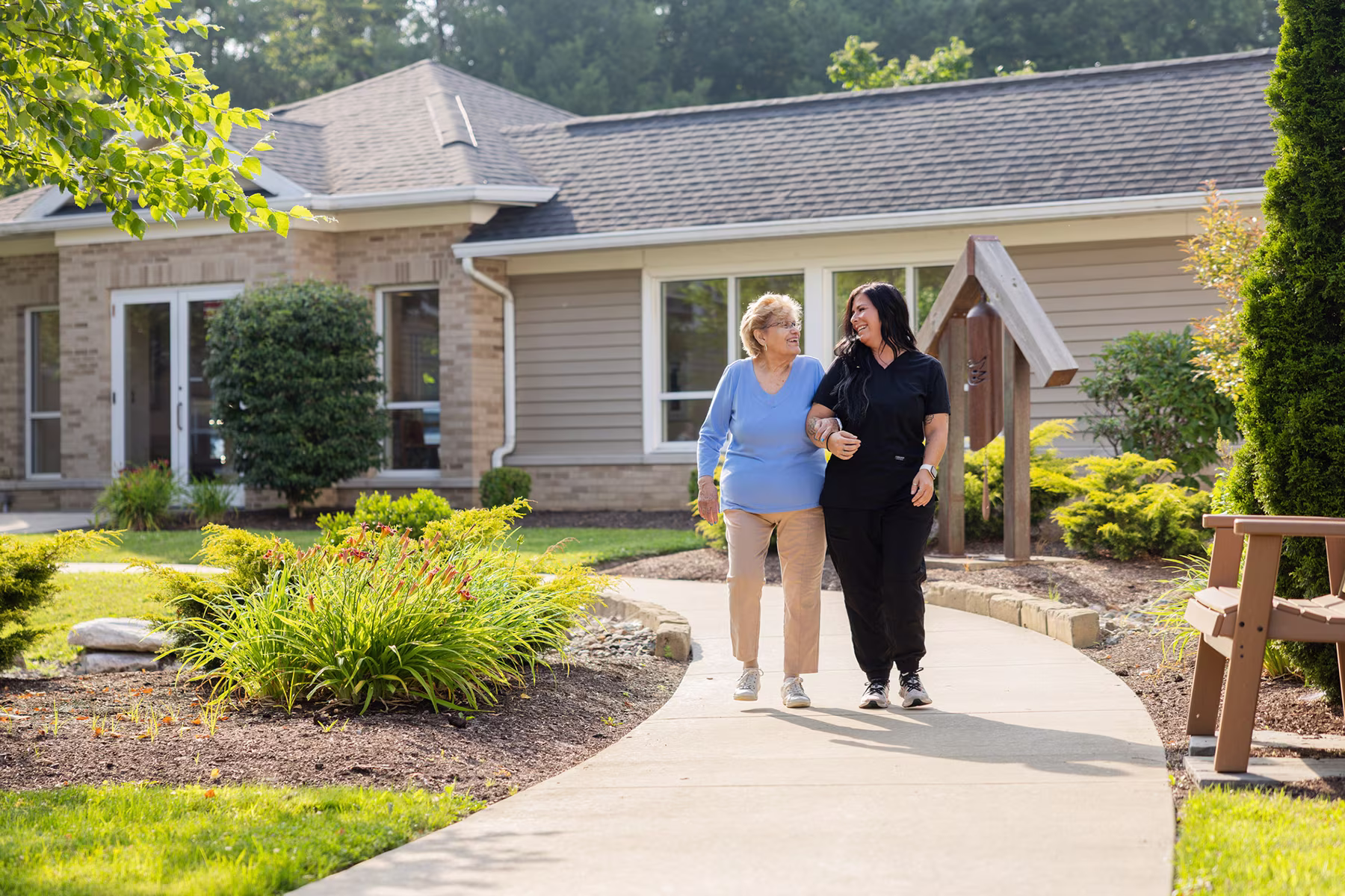 An elderly woman and a younger woman walking arm in arm on a curved garden path outside a single-story house with greenery and shrubs around them, both smiling and engaged in conversation.