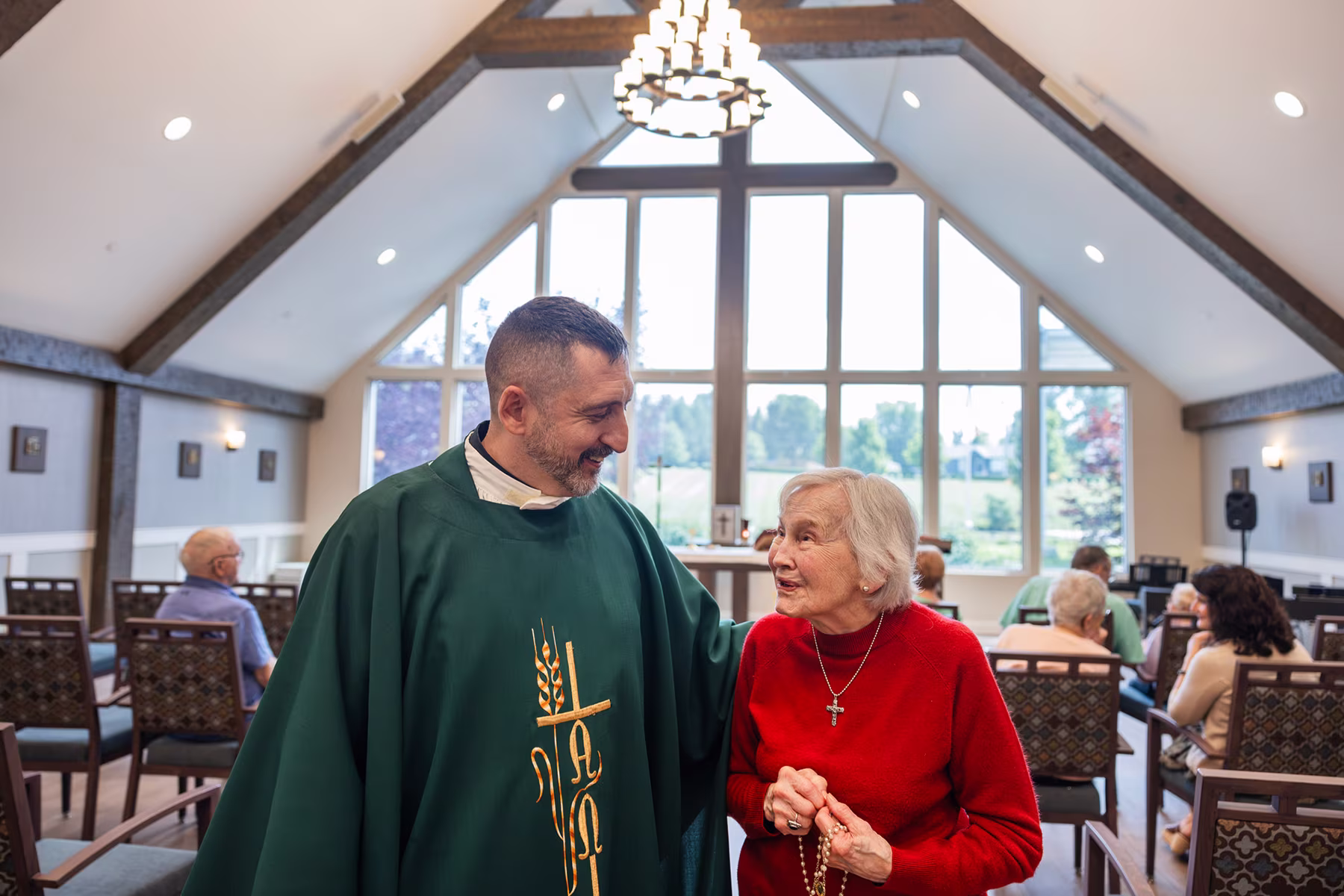 A male priest in a green robe with gold embroidery smiles and speaks with an elderly woman in a red sweater wearing a cross necklace inside a well-lit chapel with large windows and seated people.