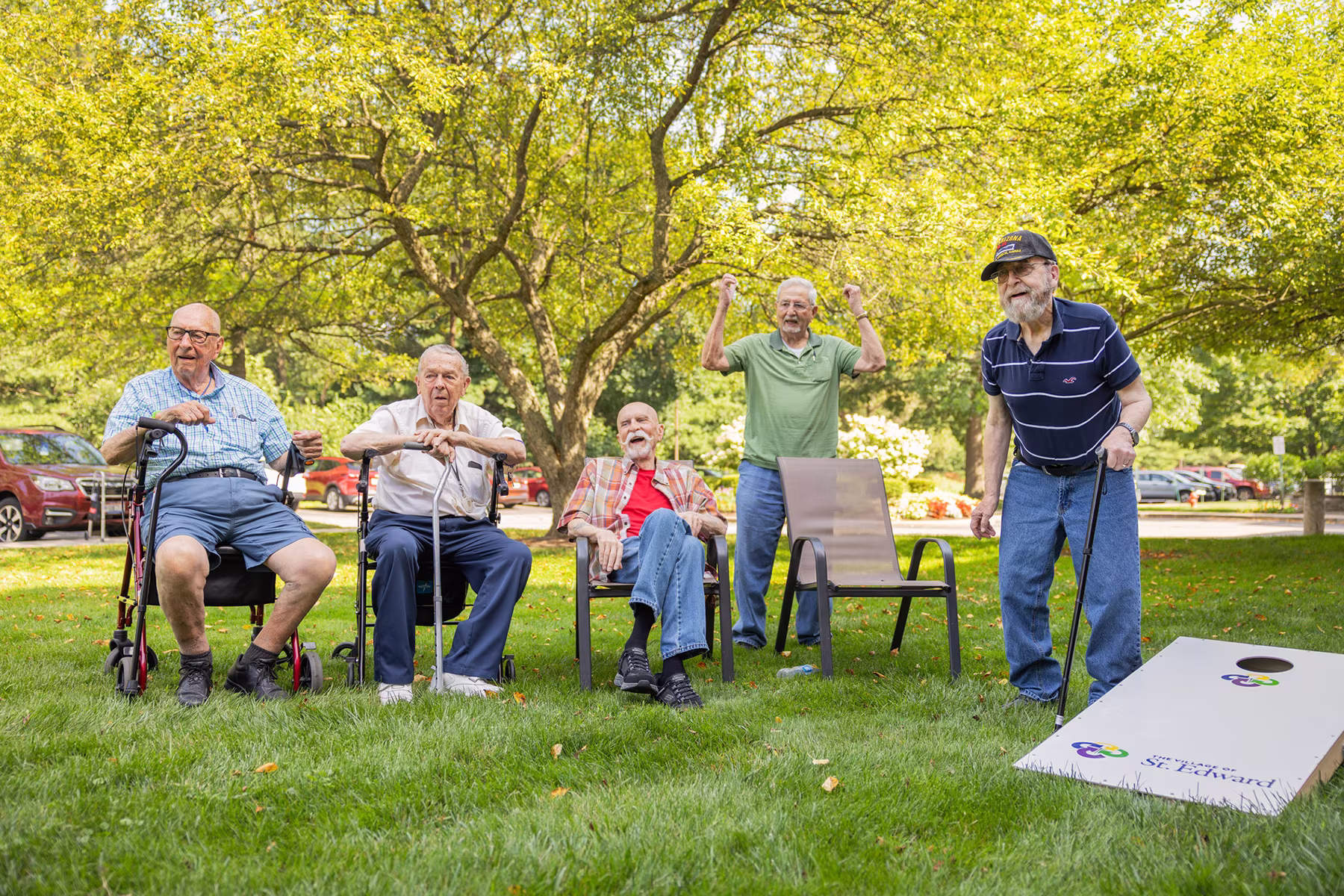 Five elderly men outdoors on grass near trees and parked cars, three seated on chairs or a walker, one standing with arms raised, and one playing cornhole with a cane, enjoying a sunny day.