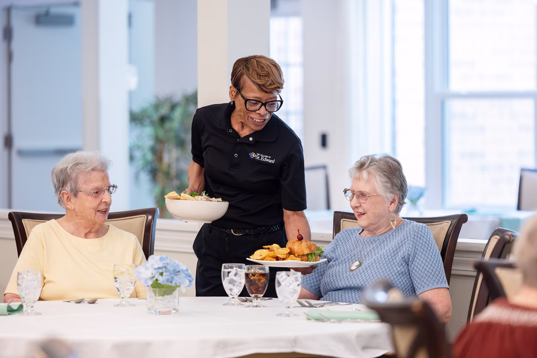 A smiling female server in a black uniform serves food to two elderly women seated at a dining table with glasses and a flower centerpiece in a well-lit room.
