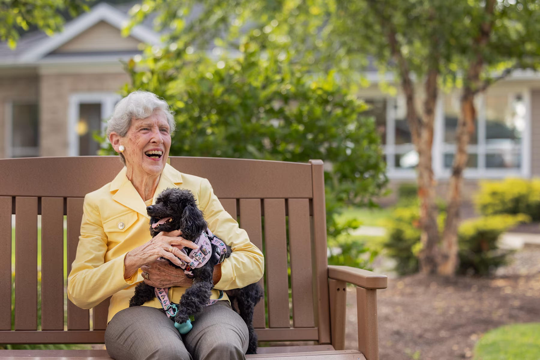 Elderly woman in a yellow jacket sitting on a wooden bench outdoors, smiling and holding a small black dog wearing a harness.