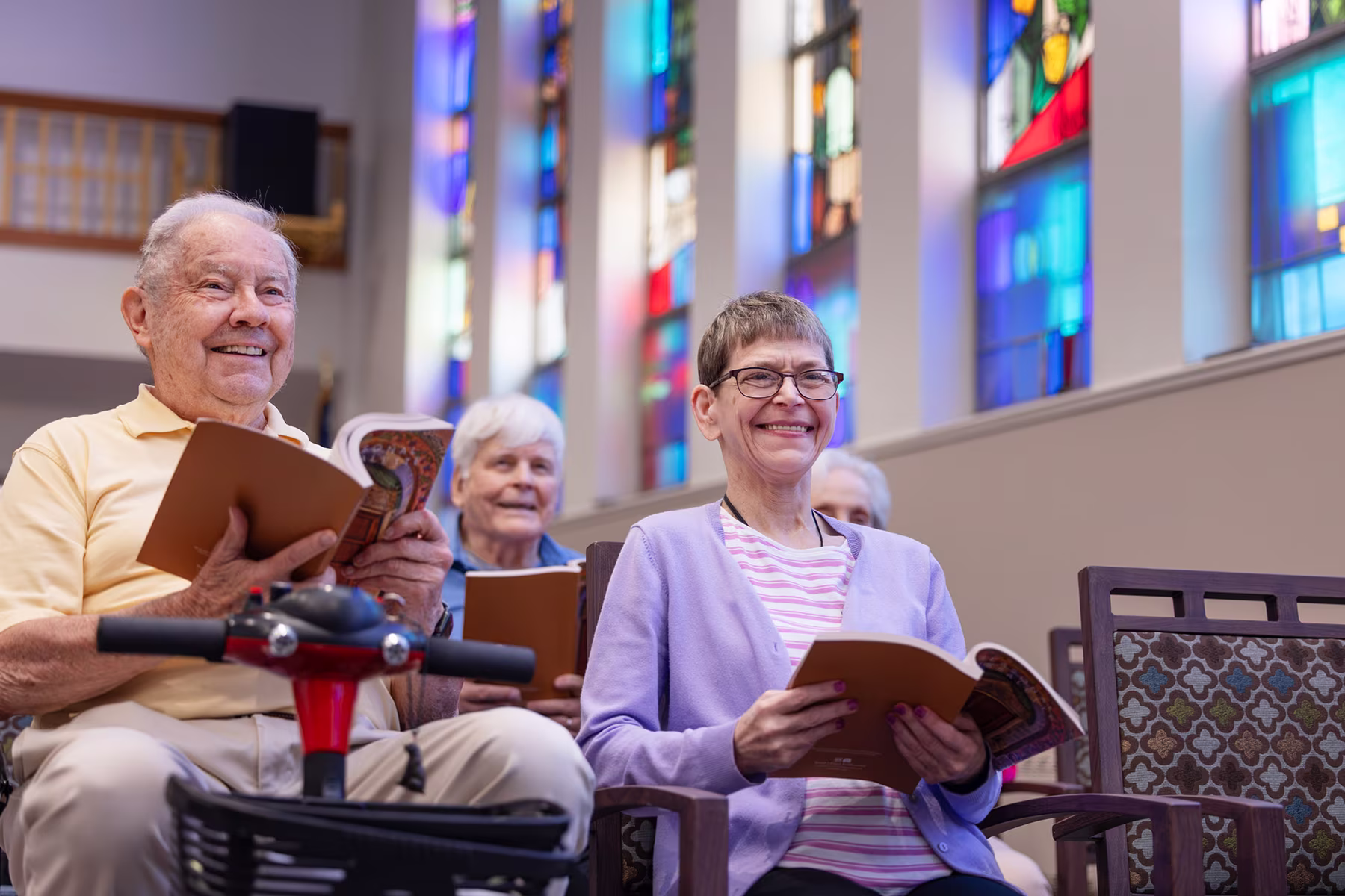 Smiling elderly people sitting in a church with stained glass windows, holding hymn books, participating in a group activity.