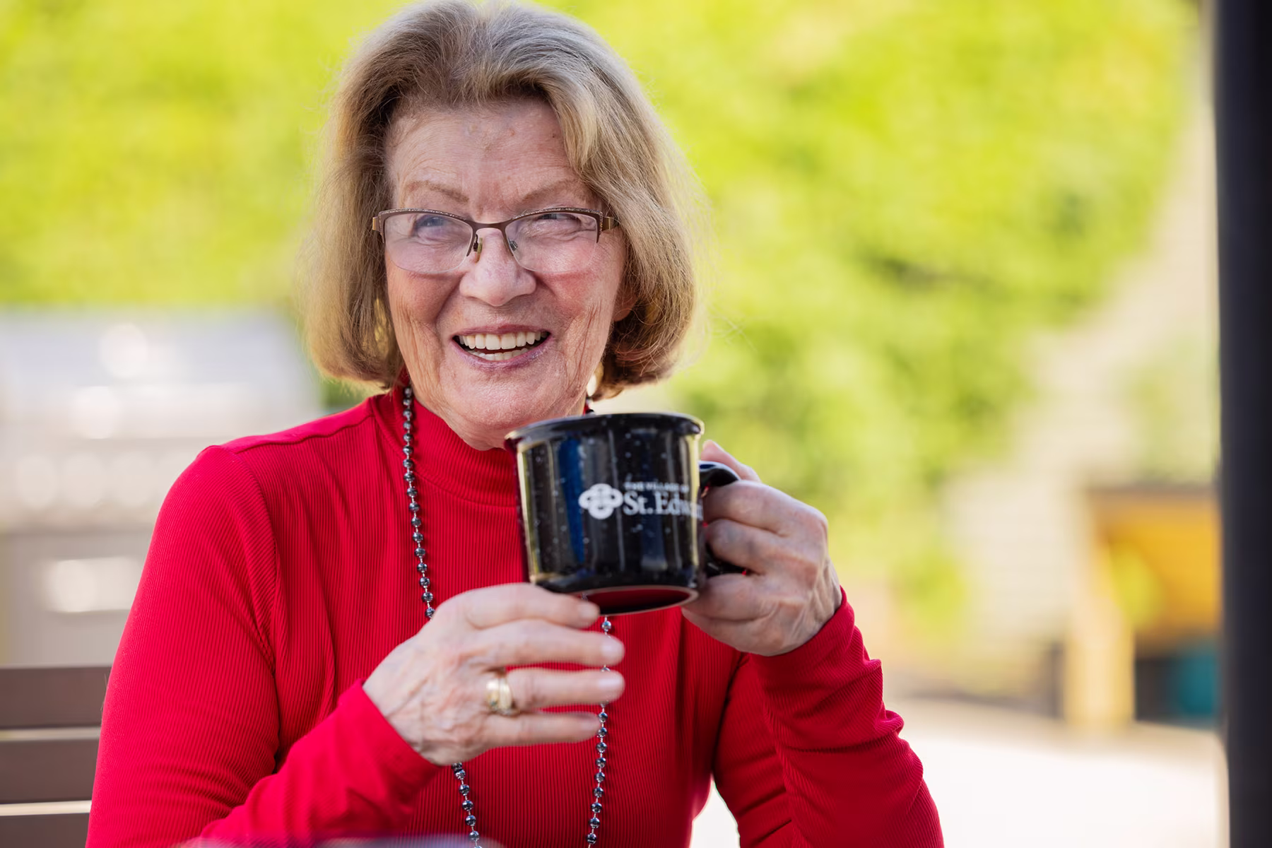 Smiling elderly woman wearing glasses and a red sweater holding a black mug that says 'St. Edna' in an outdoor setting.