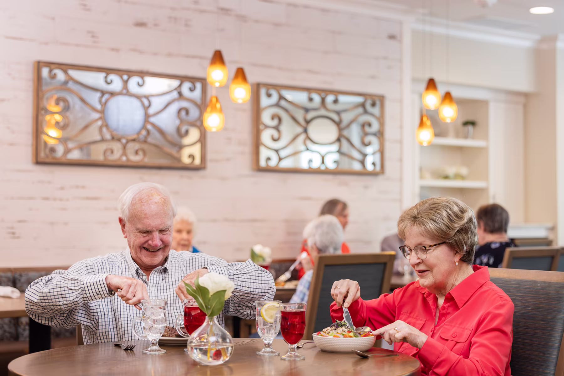 Two elderly people smiling and eating at a restaurant table, with drinks, a vase with a white flower, and decorative mirrors on the wall behind them.