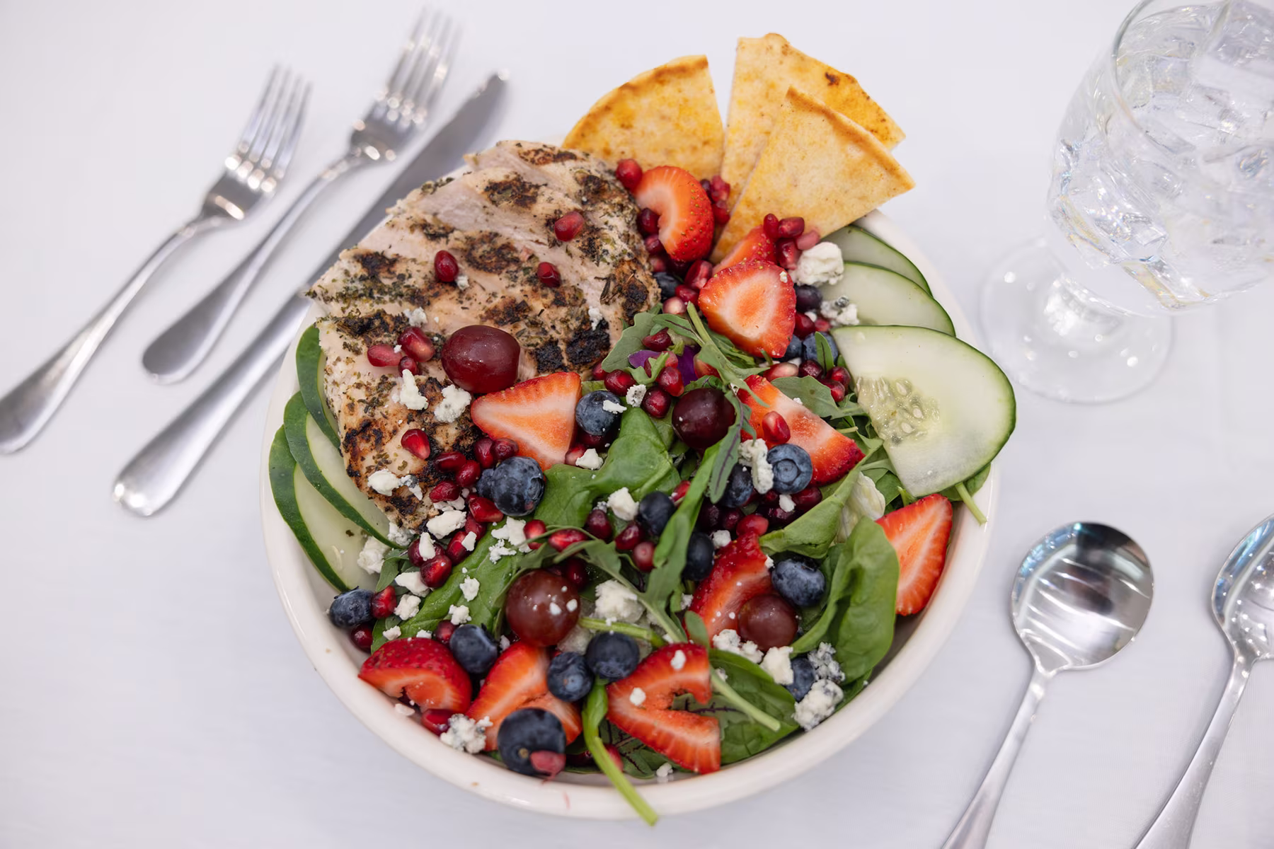 Bowl with grilled chicken breast, spinach, strawberries, blueberries, grapes, cucumber slices, pomegranate seeds, feta cheese, and tortilla chips, placed on a white tablecloth with forks and spoons nearby and a glass of water.