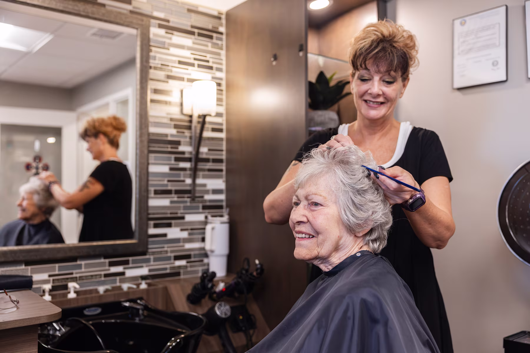Smiling hairstylist cutting the hair of an elderly woman in a salon with tiled wall and mirror reflecting them.
