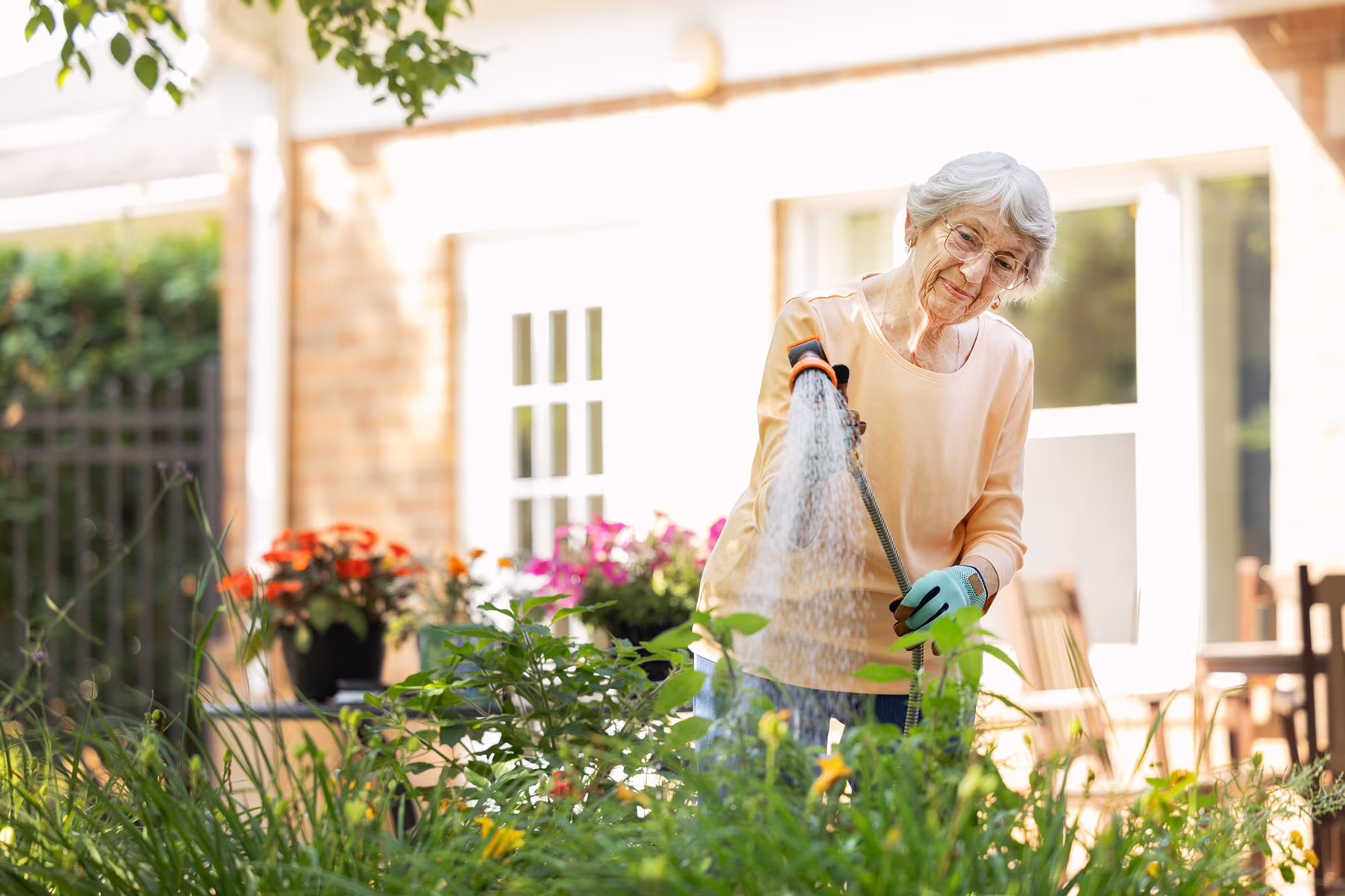 An elderly woman wearing glasses and gloves waters plants in a sunlit garden with flowers and green foliage near a house.