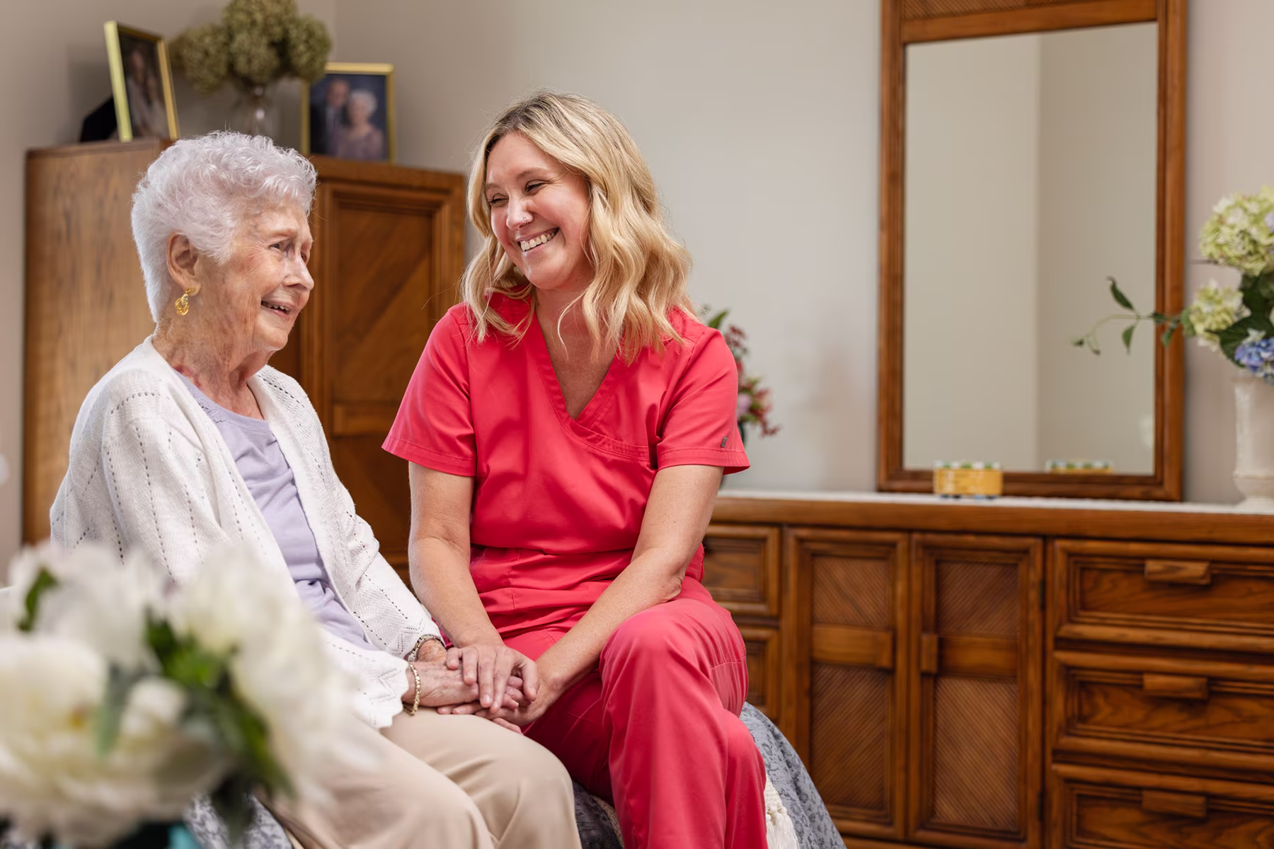 Smiling elderly woman in a white cardigan sitting beside a female caregiver in red scrubs holding hands and sharing a warm moment indoors.