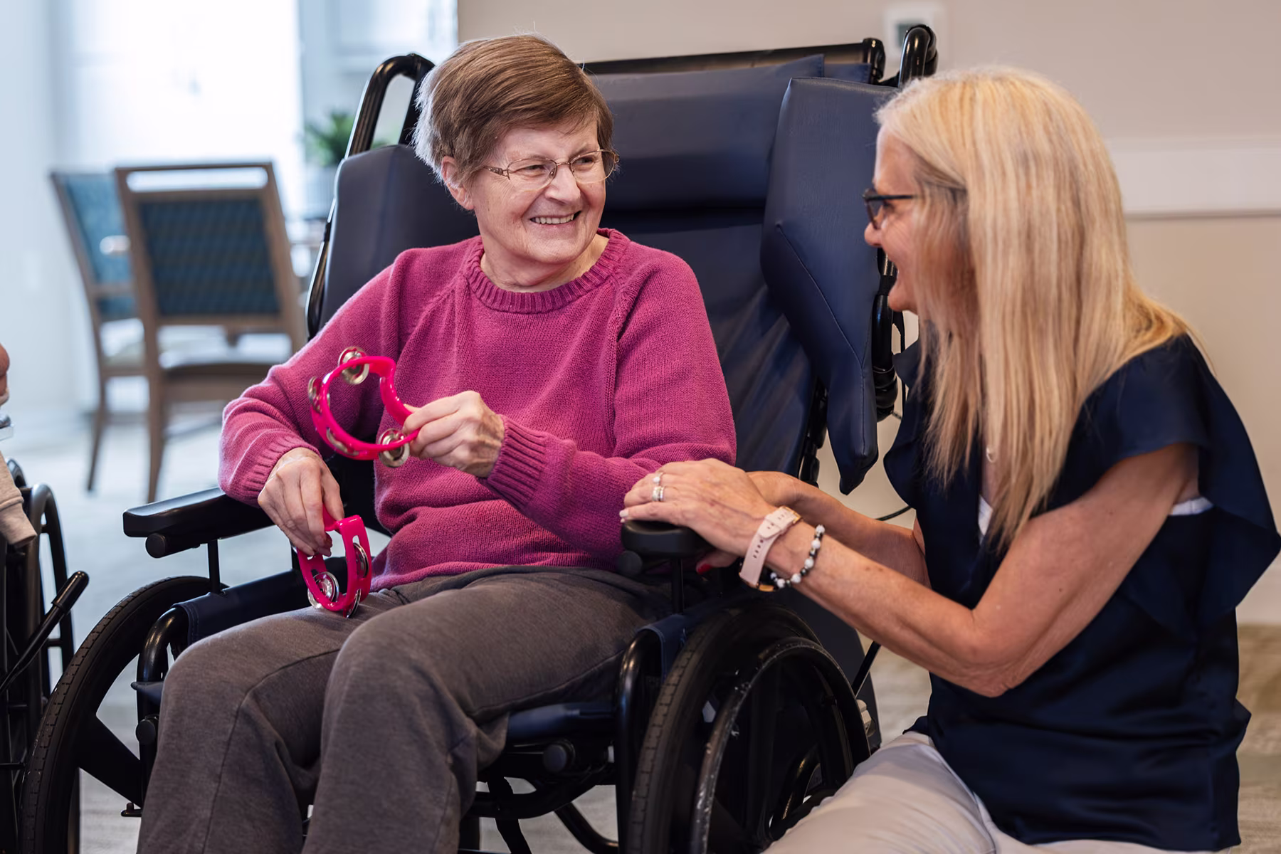 An elderly woman in a wheelchair holding pink tambourines and smiling at a blonde woman kneeling beside her who is also smiling and holding the wheelchair's armrest.