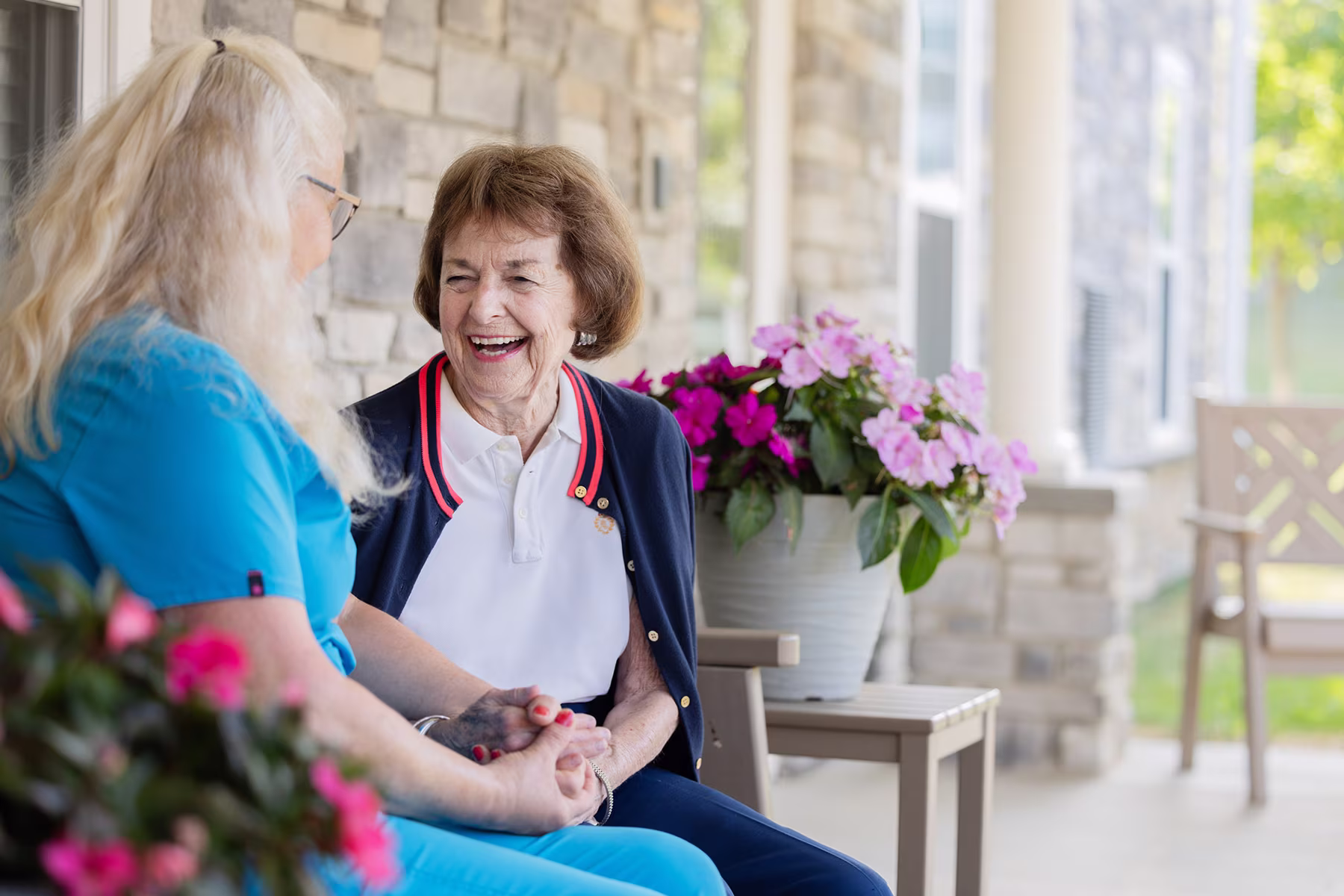 An elderly woman sitting outdoors on a porch, holding hands and smiling with a caregiver dressed in blue scrubs, surrounded by potted pink flowers and a stone wall background.