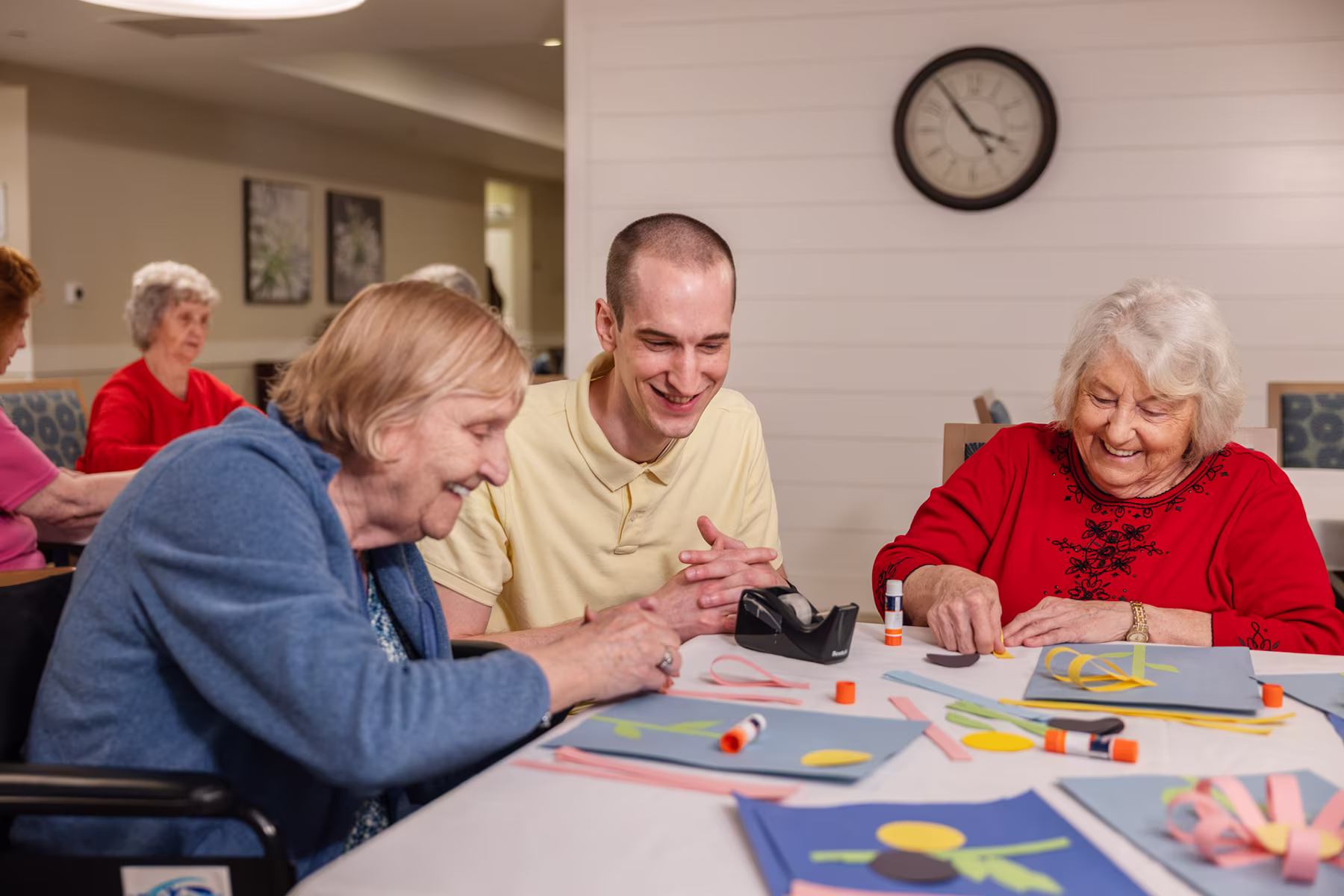 A young man and two elderly women smiling and working on colorful arts and crafts at a table, with glue sticks and paper cutouts in a cozy indoor setting with more people and a clock in the background.