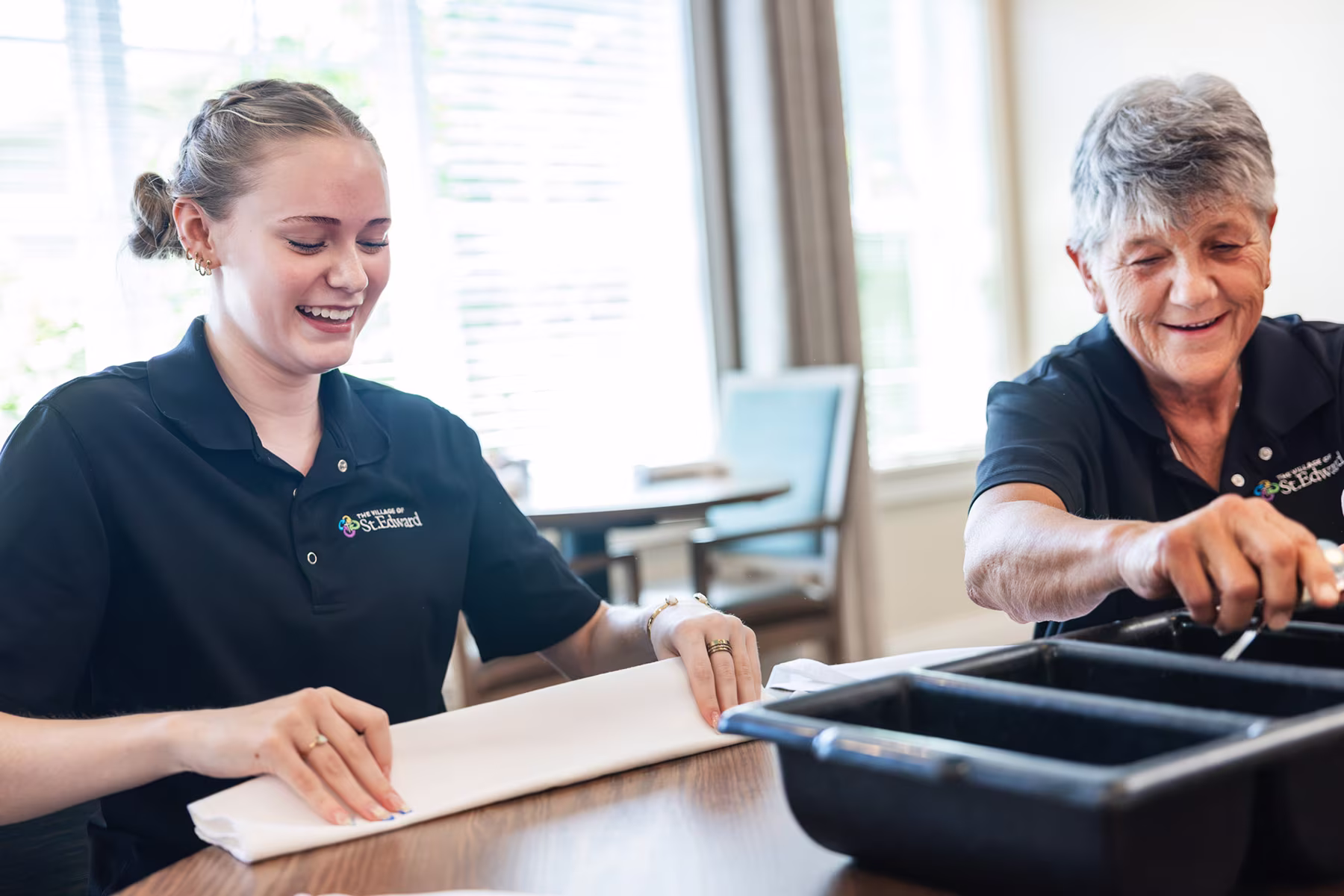 Two women in black polo shirts smiling and folding napkins at a wooden table with baking trays in front of them, inside a bright room with chairs and windows.