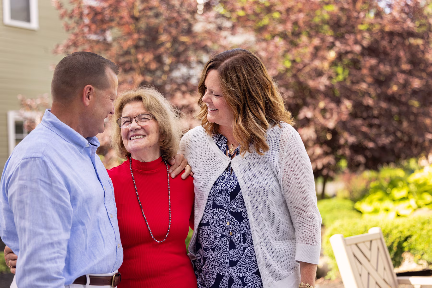Three people outdoors in a garden setting, smiling and interacting warmly; an older woman in a red sweater and glasses stands between a man in a light blue shirt and a woman in a white cardigan with patterned blue dress.