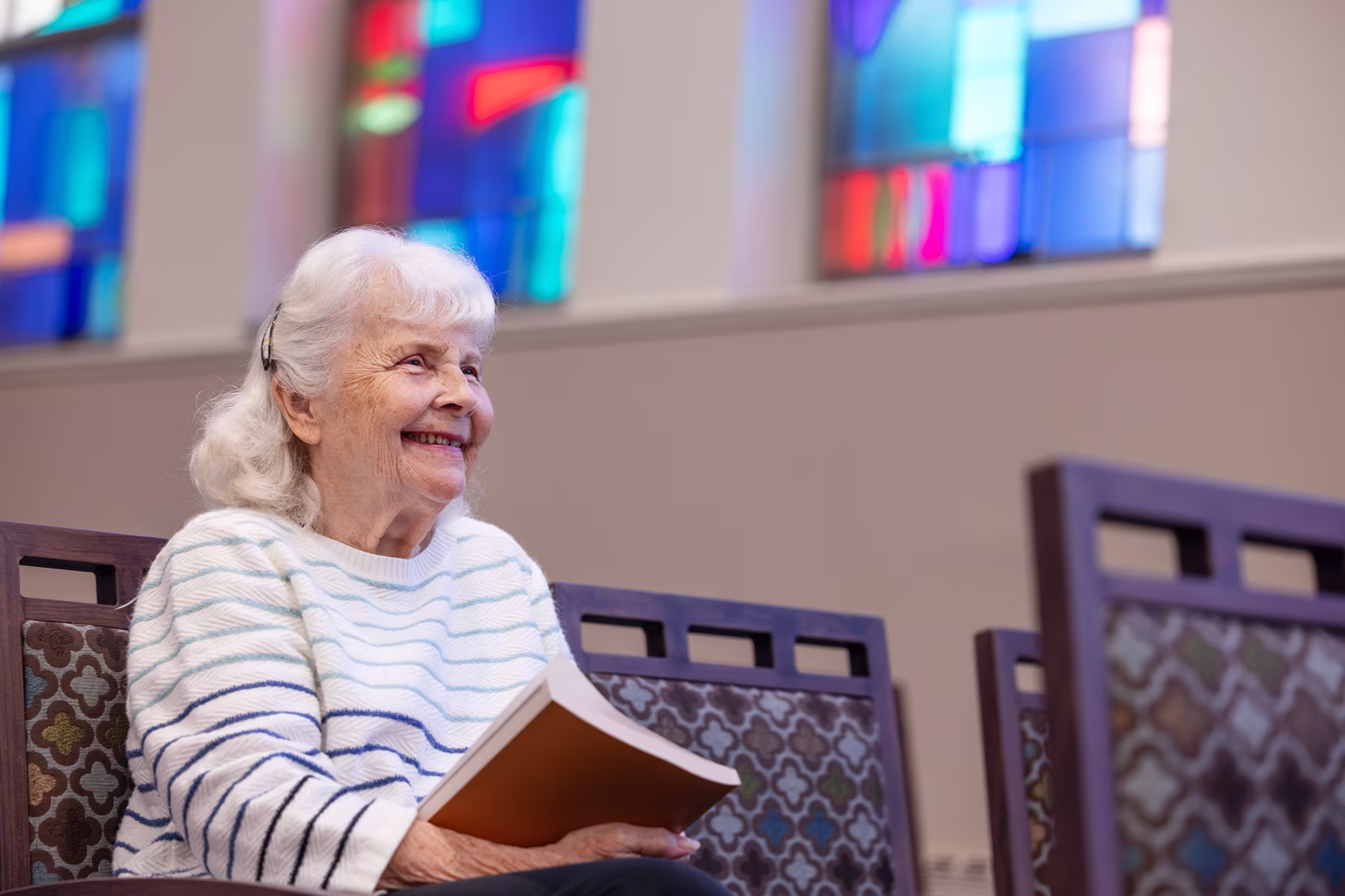 Elderly woman with white hair smiling and holding a book, sitting on a patterned chair inside a building with colorful stained glass windows in the background.