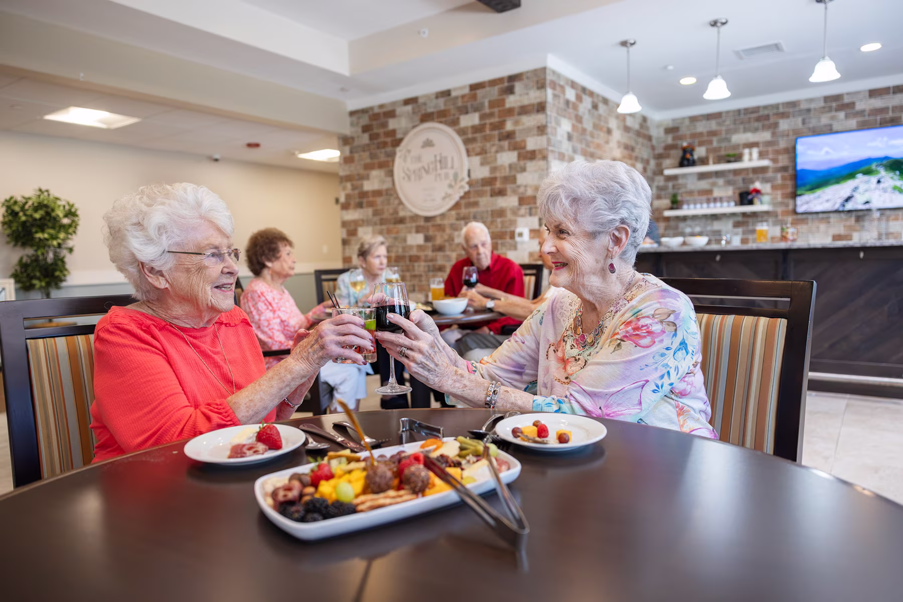 Two elderly women smiling and clinking glasses of red wine across a dining table with plates of food and a tray of fruit and snacks in a bright, modern dining room with other seniors in the background.