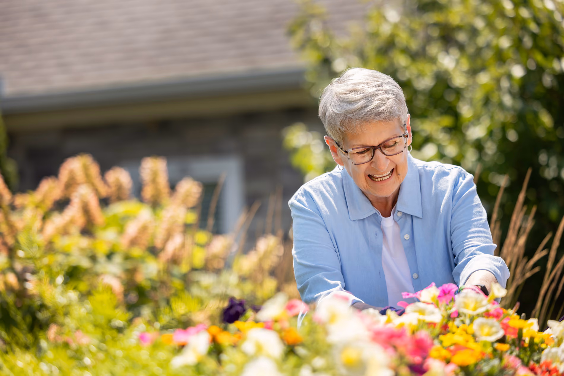 Smiling elderly woman with glasses and short gray hair tends to colorful flowers in a garden outside a house.