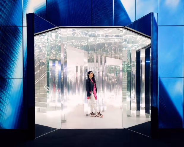 WOMAN STANDS AMIDST MIRRORS IN MIAMI’S IMMERSIVE ART MUSEUM CALLED SUPERBLUE IN INSTALLATION CALLED THE FOREST OF US
