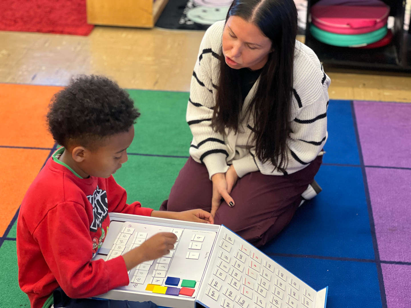 Teacher guides young student through a number activity on colorful classroom carpet