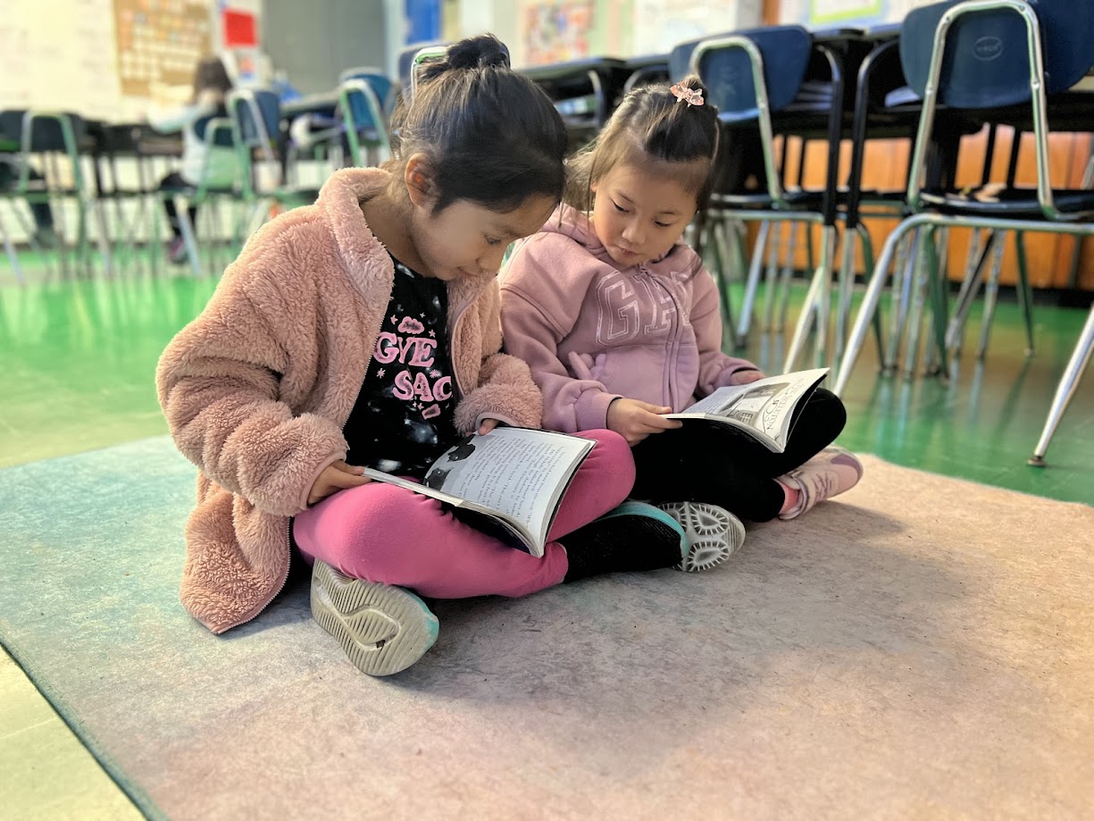 Two young students sitting on classroom floor reading a book together