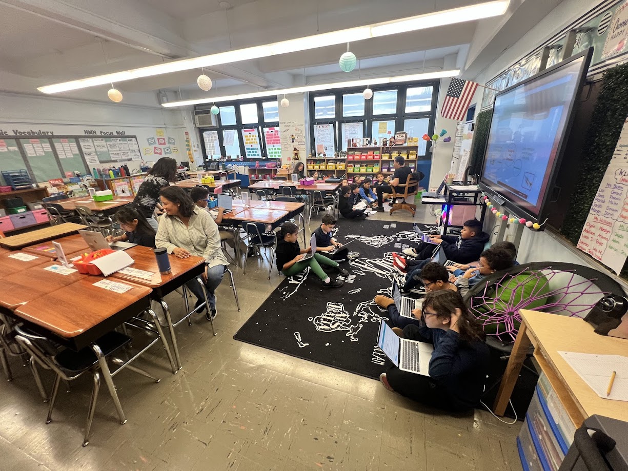 Students and teacher working on laptops in a busy elementary classroom