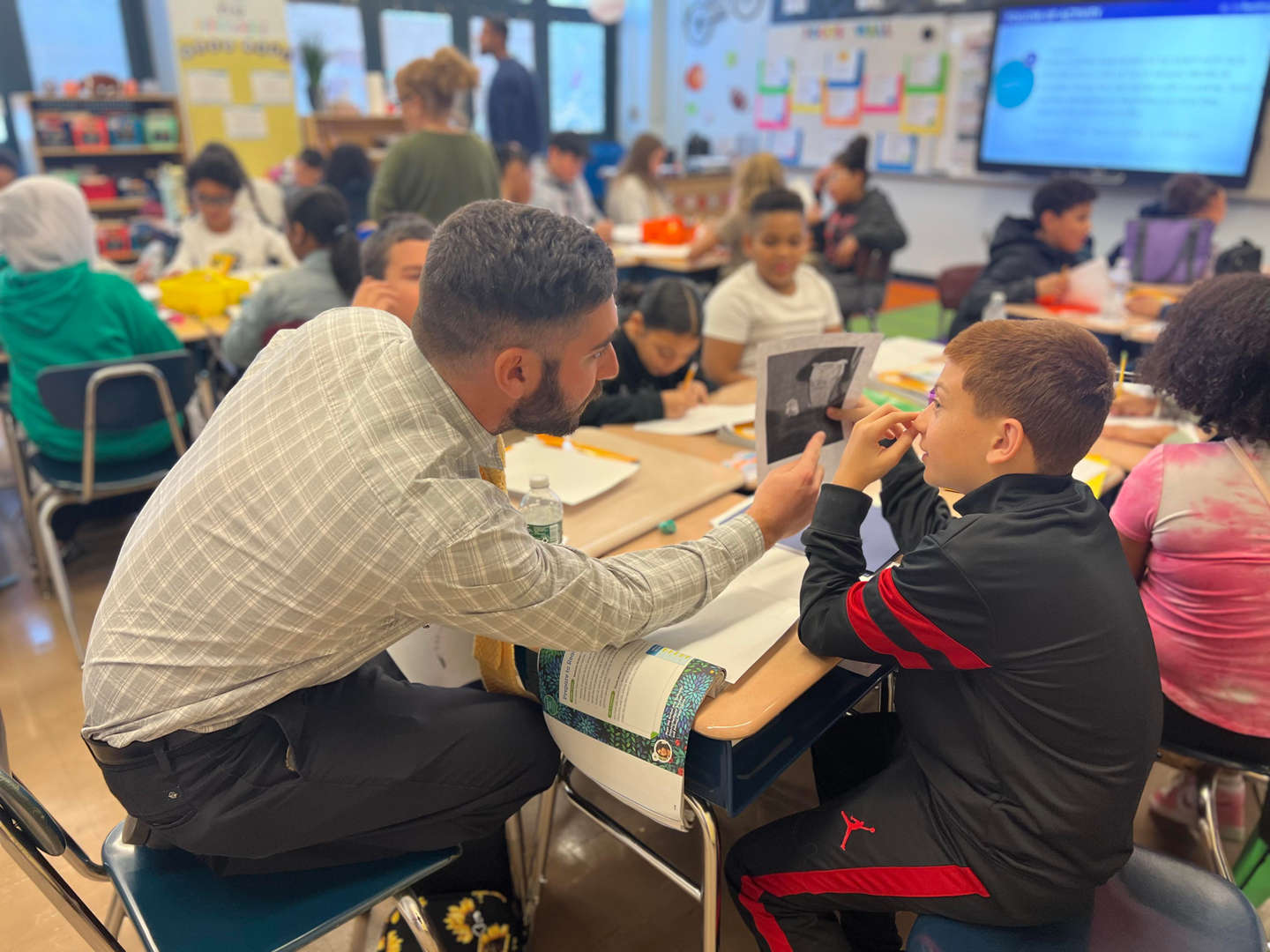 Teacher kneels beside student desk providing one-on-one reading support