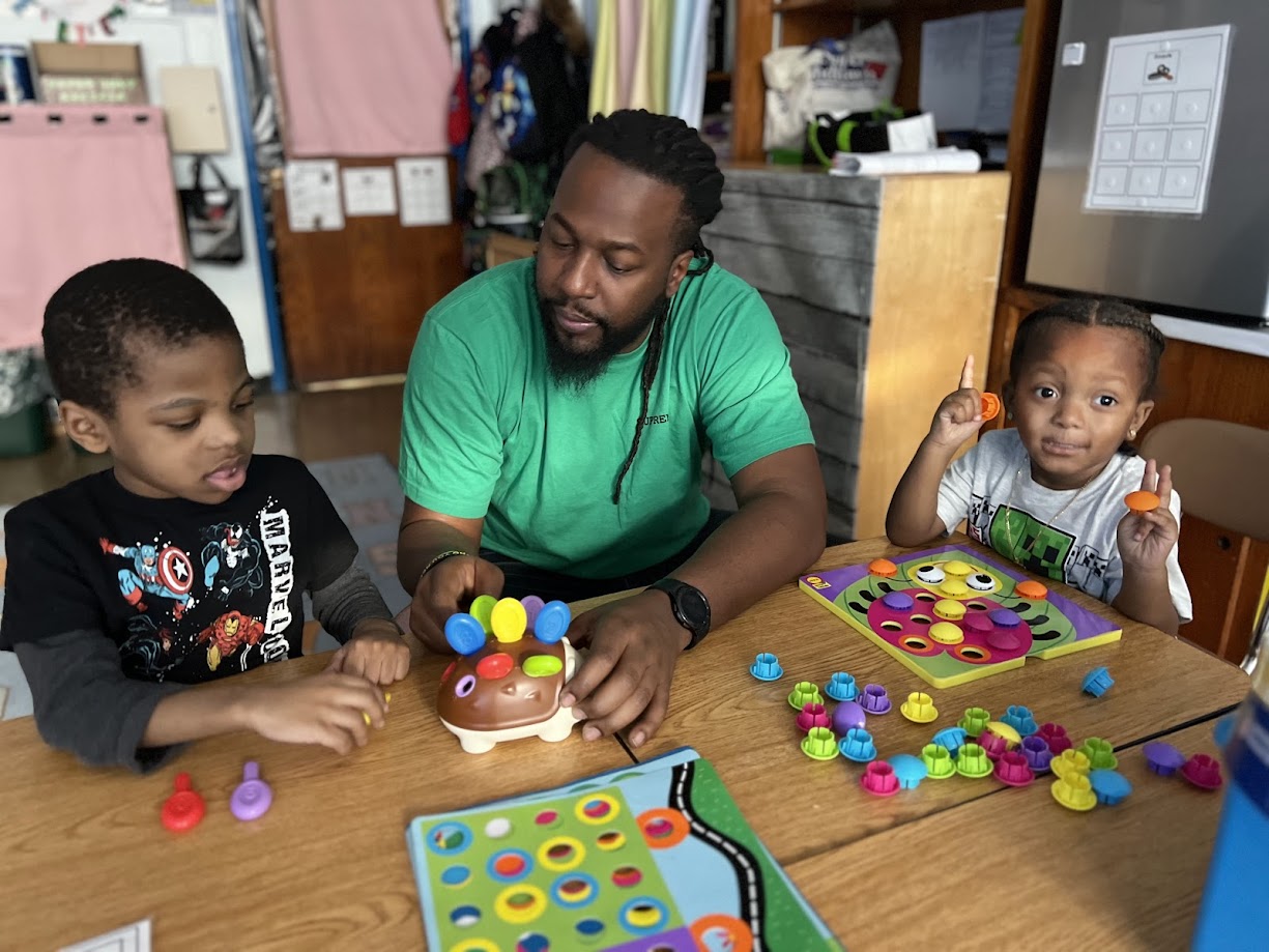 Adult and two young children playing colorful sorting games at classroom table