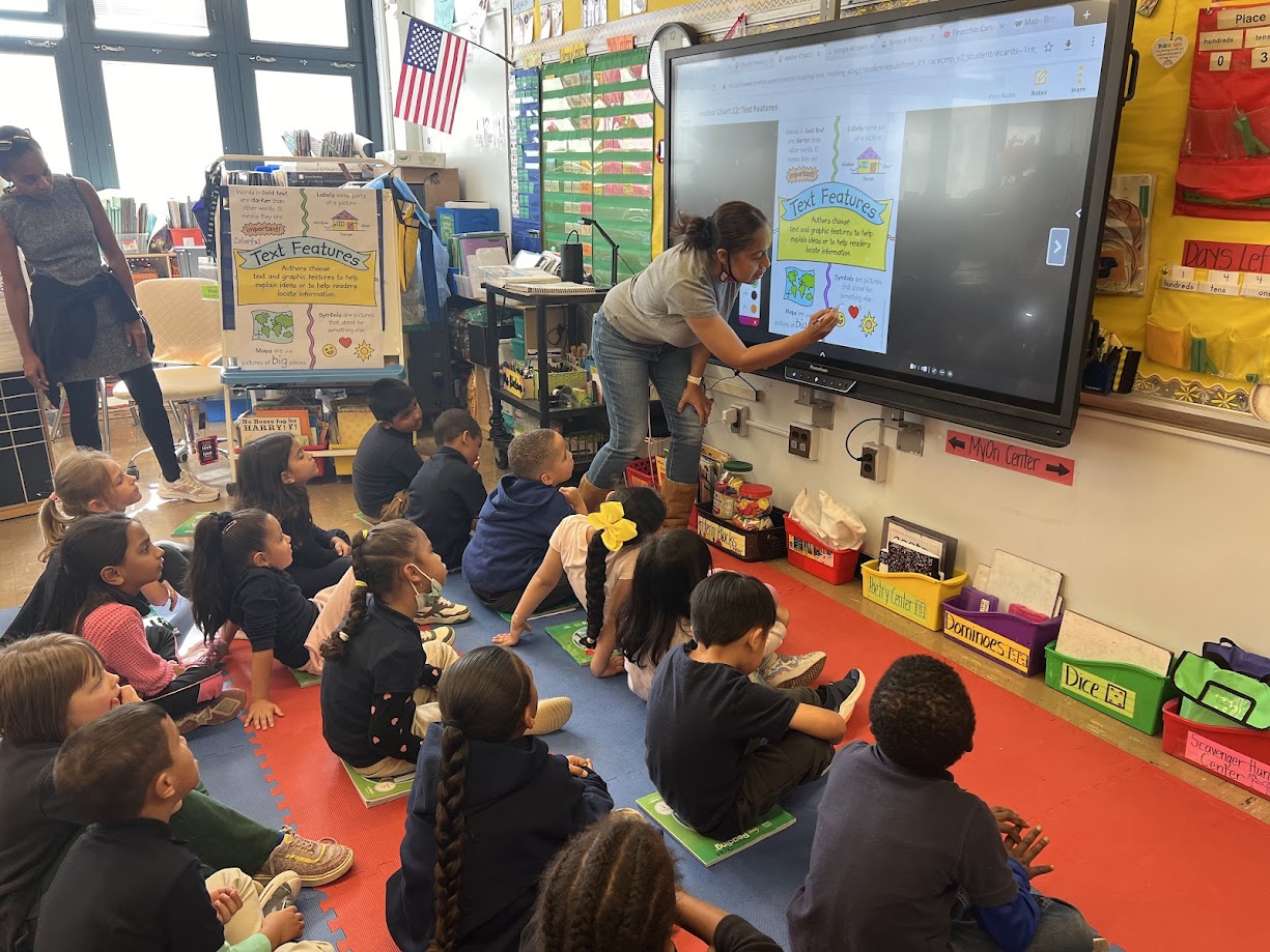 Teacher points to smartboard while students sit on classroom floor