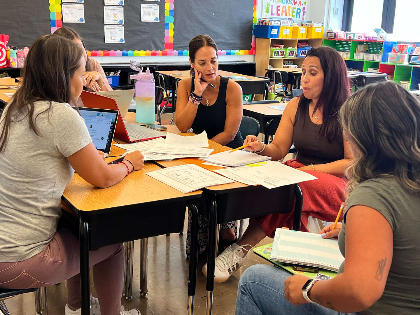 Teachers collaborating around desks with papers and laptops in classroom