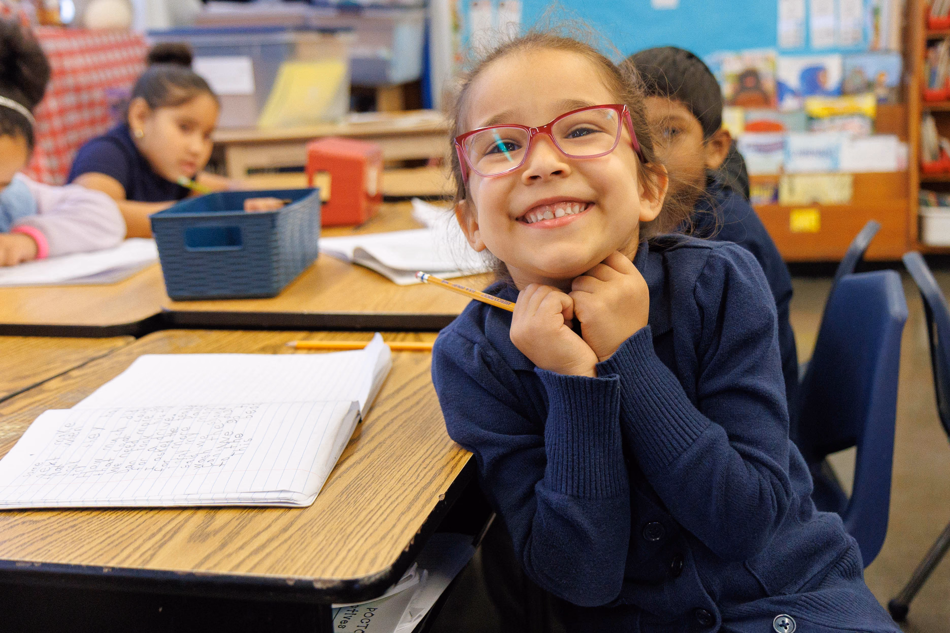 Smiling young student with glasses at classroom desk holding pencil