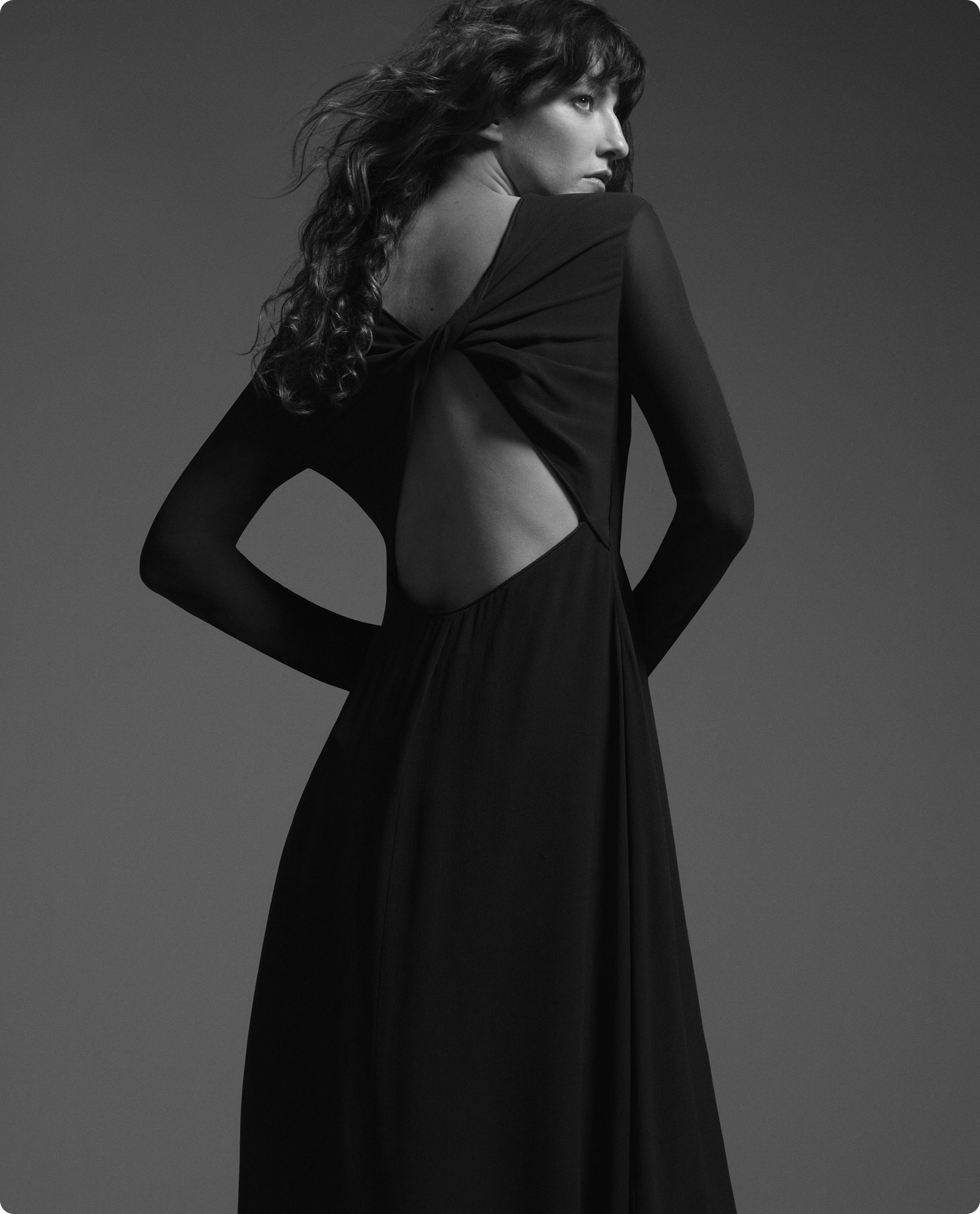 Modelo de costas em vestido preto longo com recorte nas costas, sobre fundo cinza