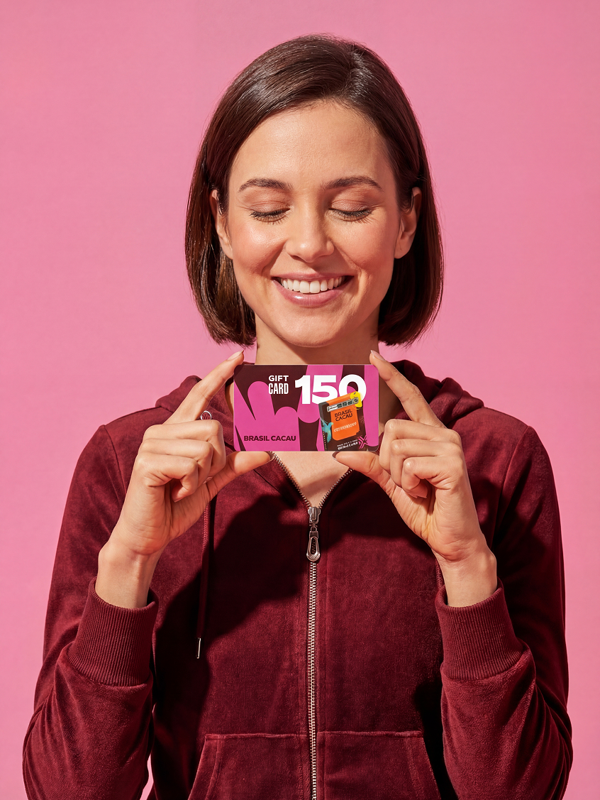 Mulher sorridente segurando cartão-presente Brasil Cacau, sobre fundo rosa