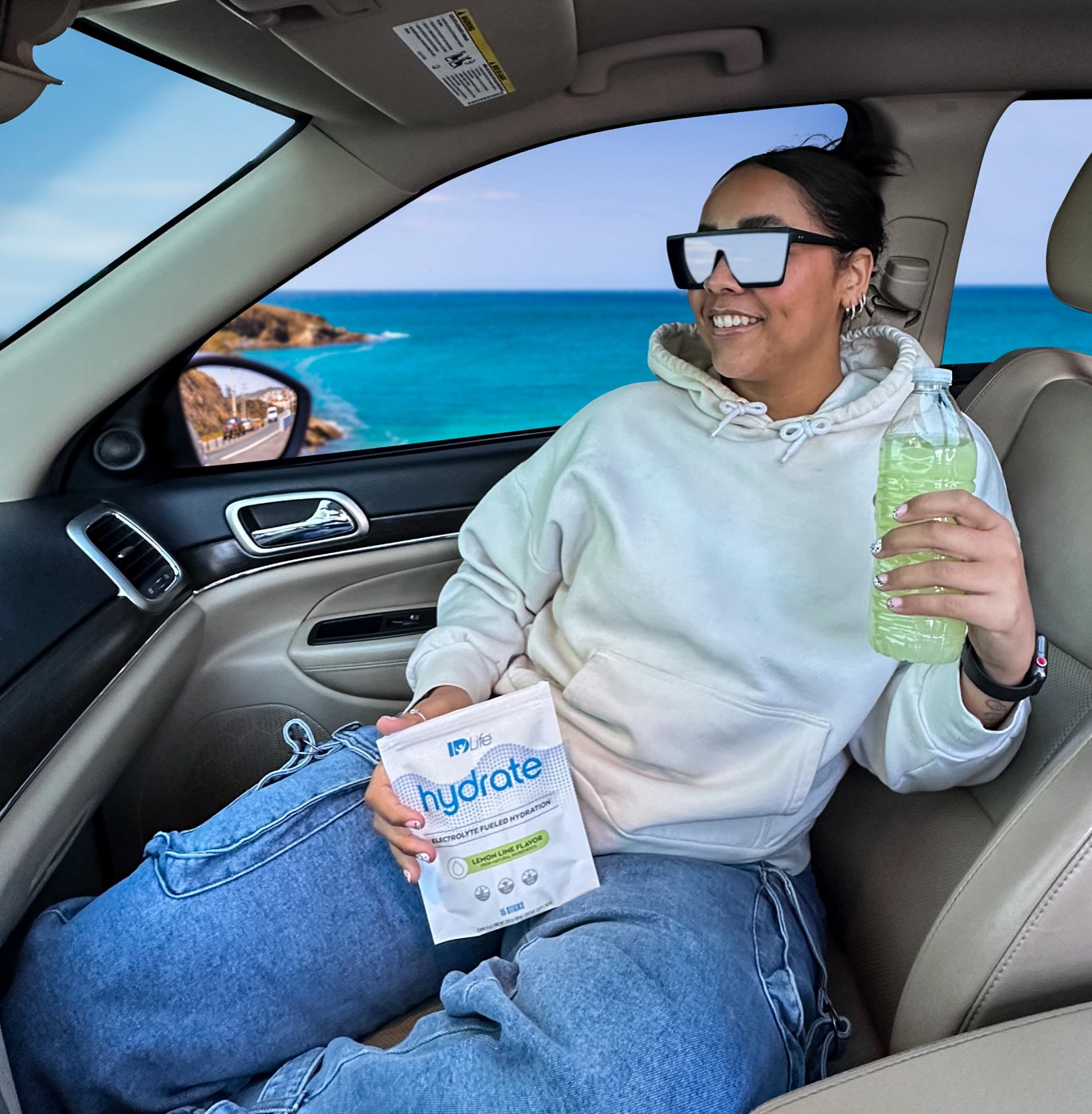A woman in the passenger seat holding a Hydrate Lemon Lime bag and a bottle, ocean out the window