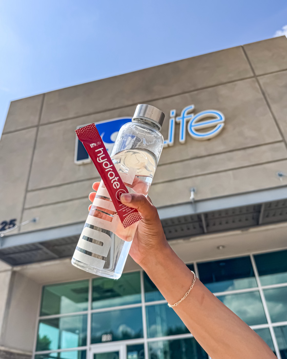 A hand holding a bottle and a red Hydrate stick up in front of the IDLife building sign
