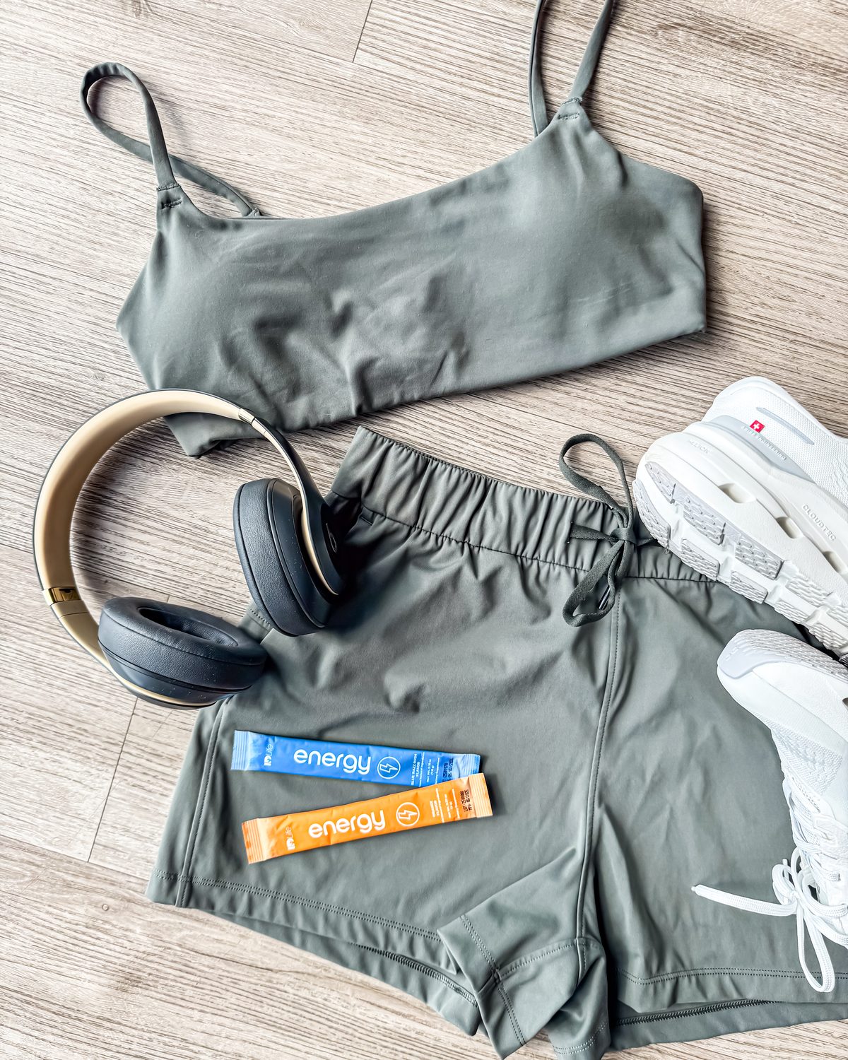 A flat lay of olive workout shorts and top, headphones and white sneakers, with a Blue Razz Rush and an Orange Energy stick on the shorts