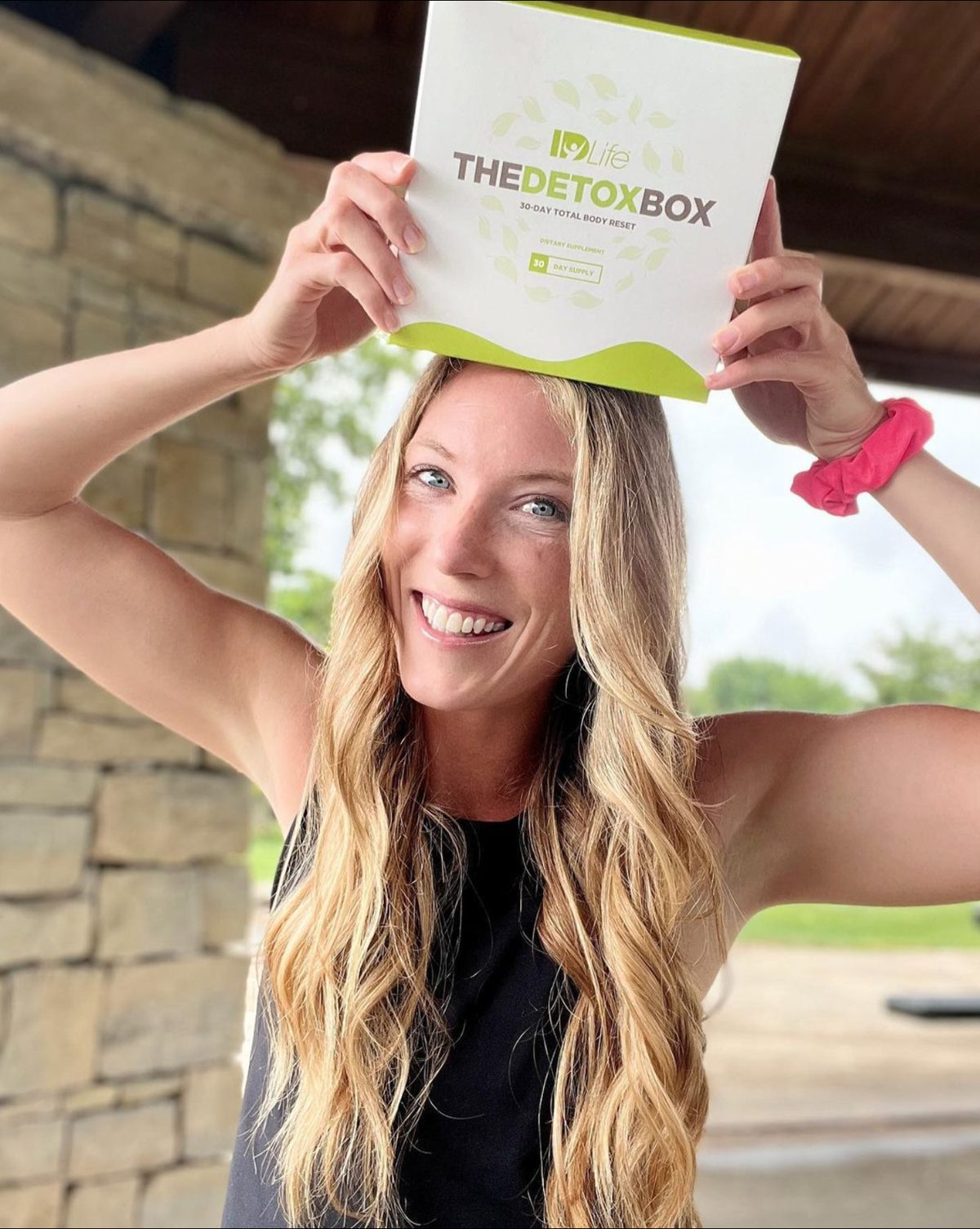 A smiling woman outdoors holding The Detox Box above her head with both hands
