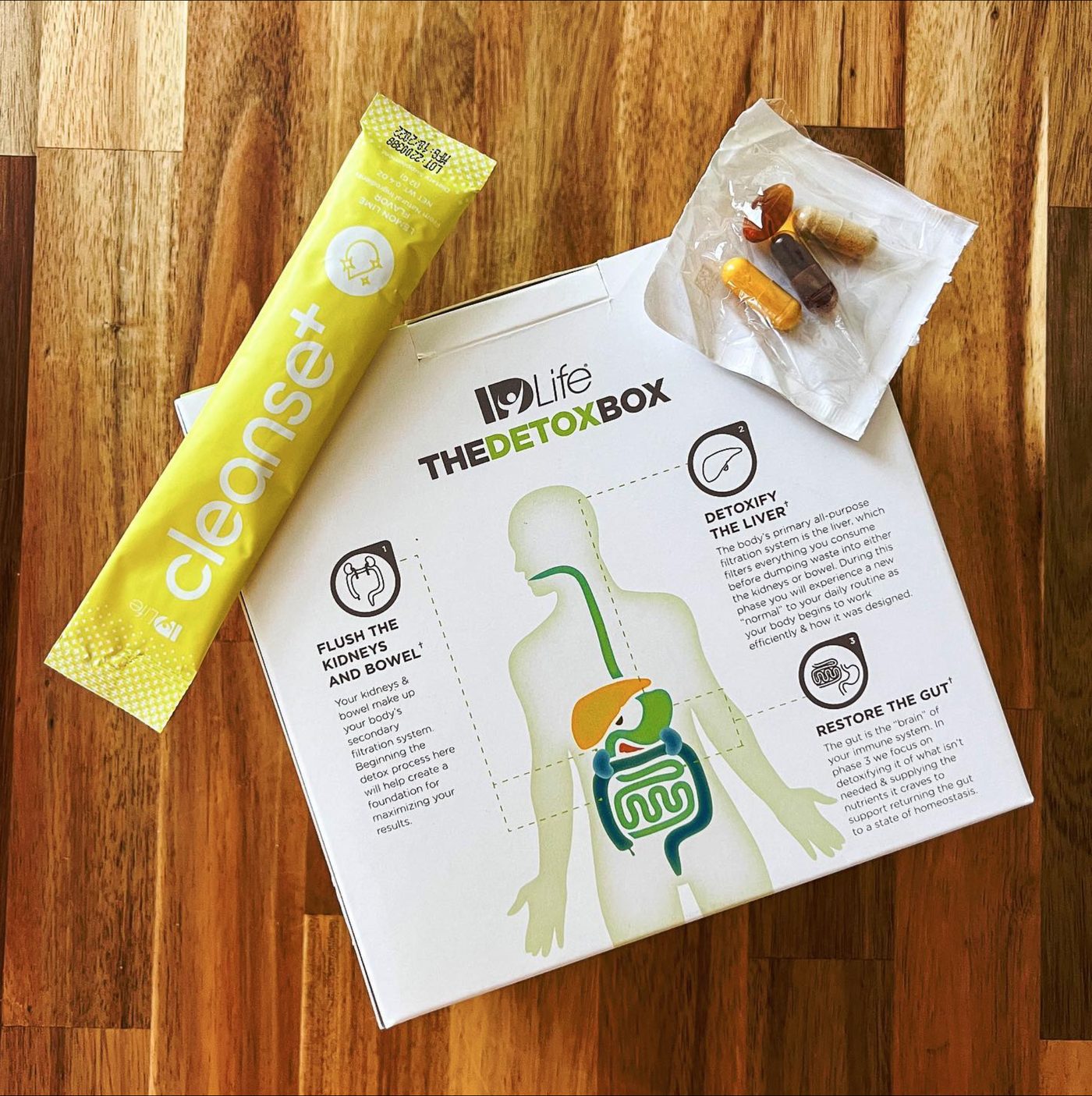 A flat lay on a wooden table: the back of The Detox Box with its three-phase diagram, a Cleanse+ Lemon-Lime stick and a torn-open pack of four capsules