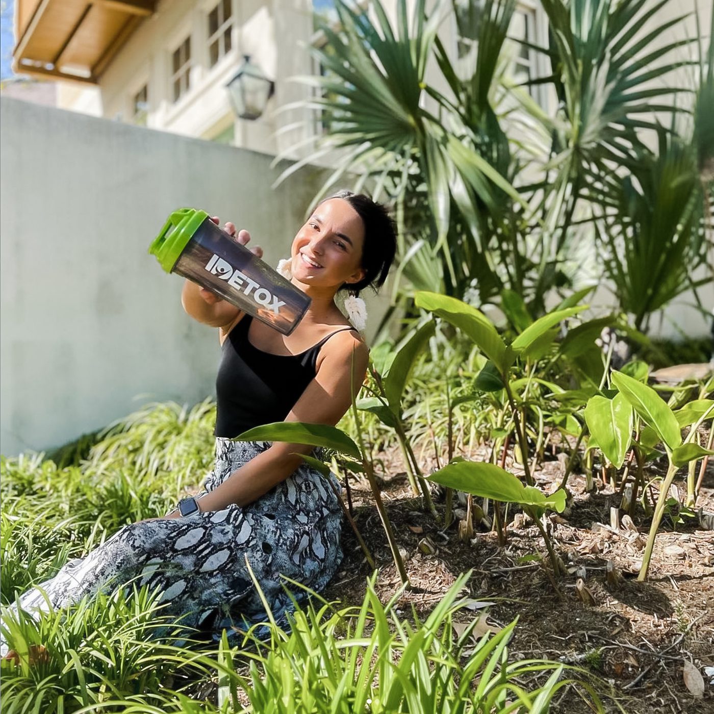 A woman sitting in a sunny garden holding up a green IDLife Detox shaker