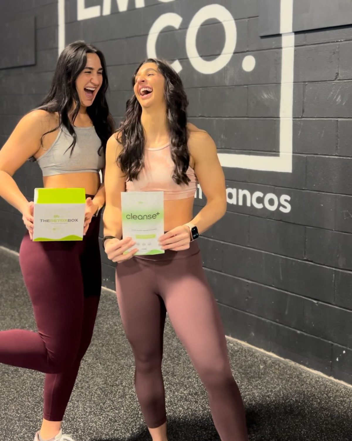 Two friends laughing in a gym, one holding The Detox Box and the other a bag of Cleanse+