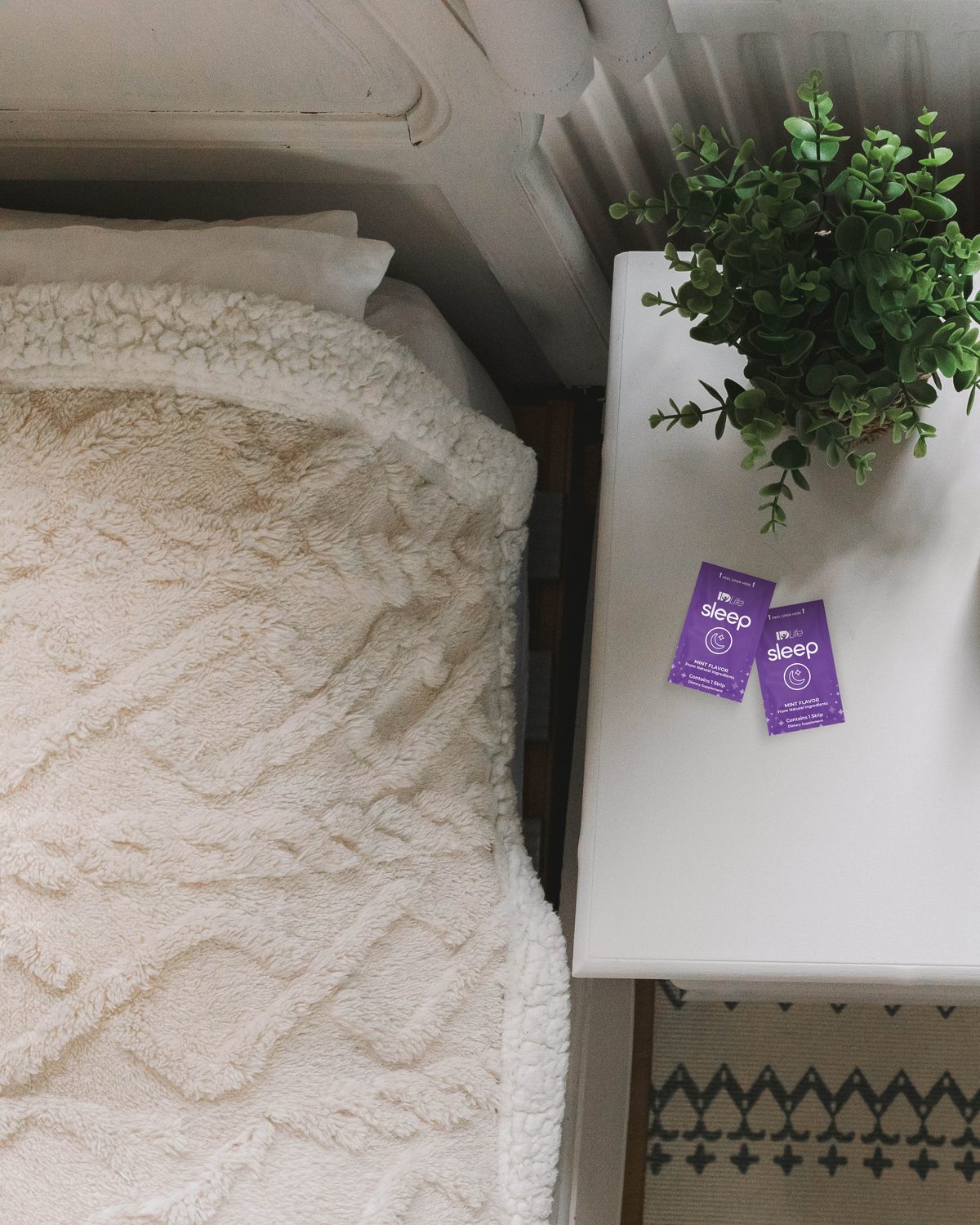 Overhead view of a bed with a cream sherpa blanket beside a white nightstand holding a green plant and two IDLife Sleep strips