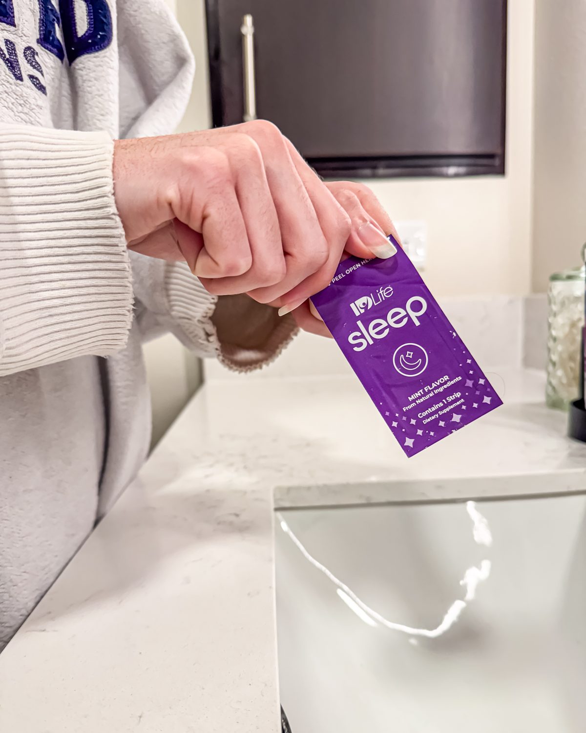 Hands in a cream knit sweater tearing open an IDLife Sleep strip pack over a bathroom counter