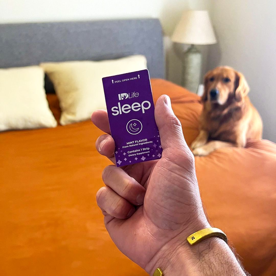 A hand holding up an IDLife Sleep strip pack in a bedroom, with a golden retriever lying on the orange bedspread behind it